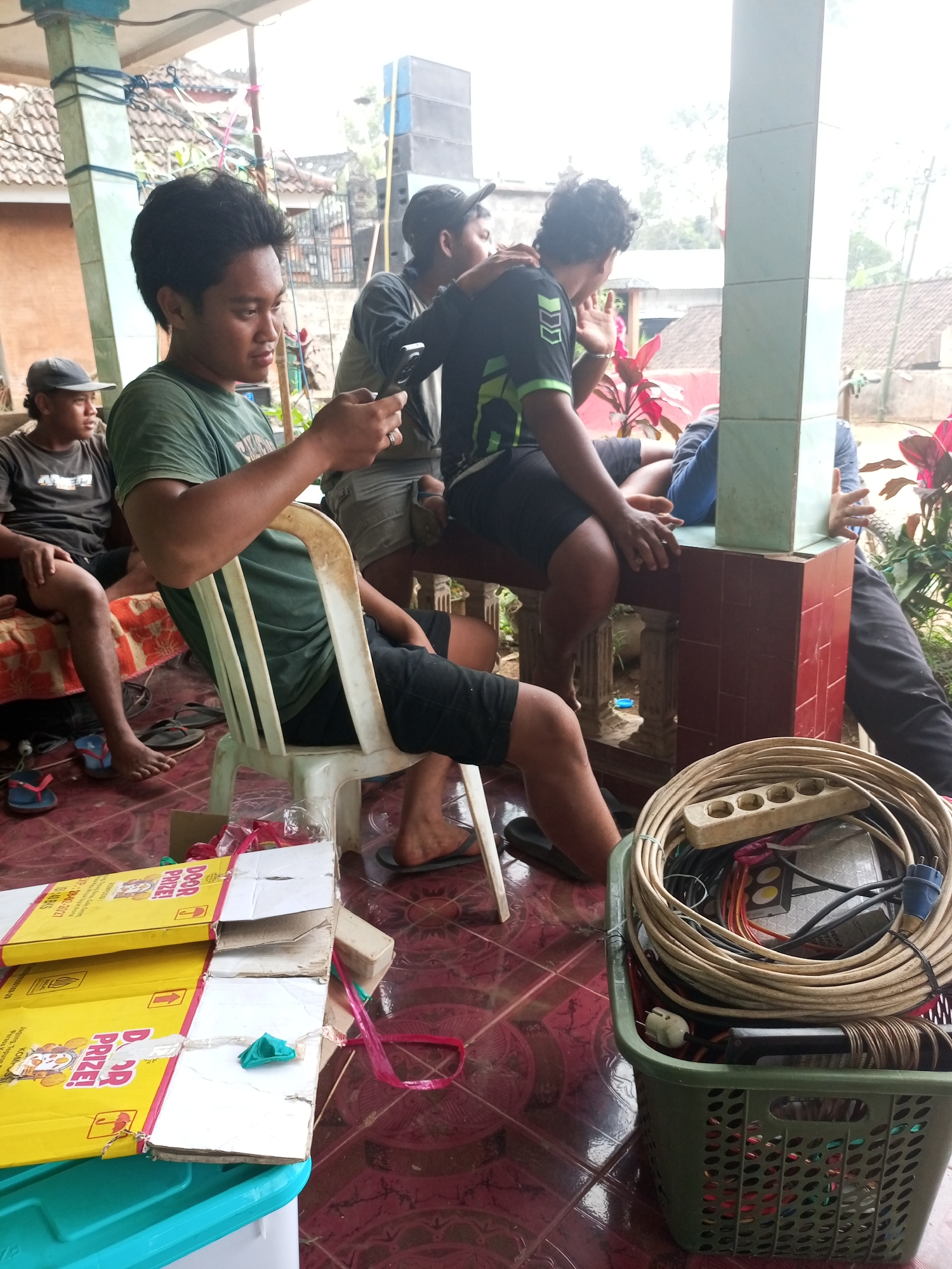 In Wonoagung, Indonesia, a group of men are gathered under a covered outdoor area. The central figure, a young man in a green t-shirt, is seated on a white plastic chair and appears to be looking at his mobile phone. To his left, another man in a grey t-shirt and cap is also seated. Further back, two other men are visible, one wearing a black t-shirt with green accents and the other in a blue shirt. The environment includes a tiled floor, a red brick and tiled railing, and exposed structural elements of the building. In the foreground, on the floor, is a green plastic basket filled with coiled electrical cords and power strips, along with some yellow cardboard boxes. The lighting suggests it is daytime, with ambient light filtering through the open sides of the covered area.