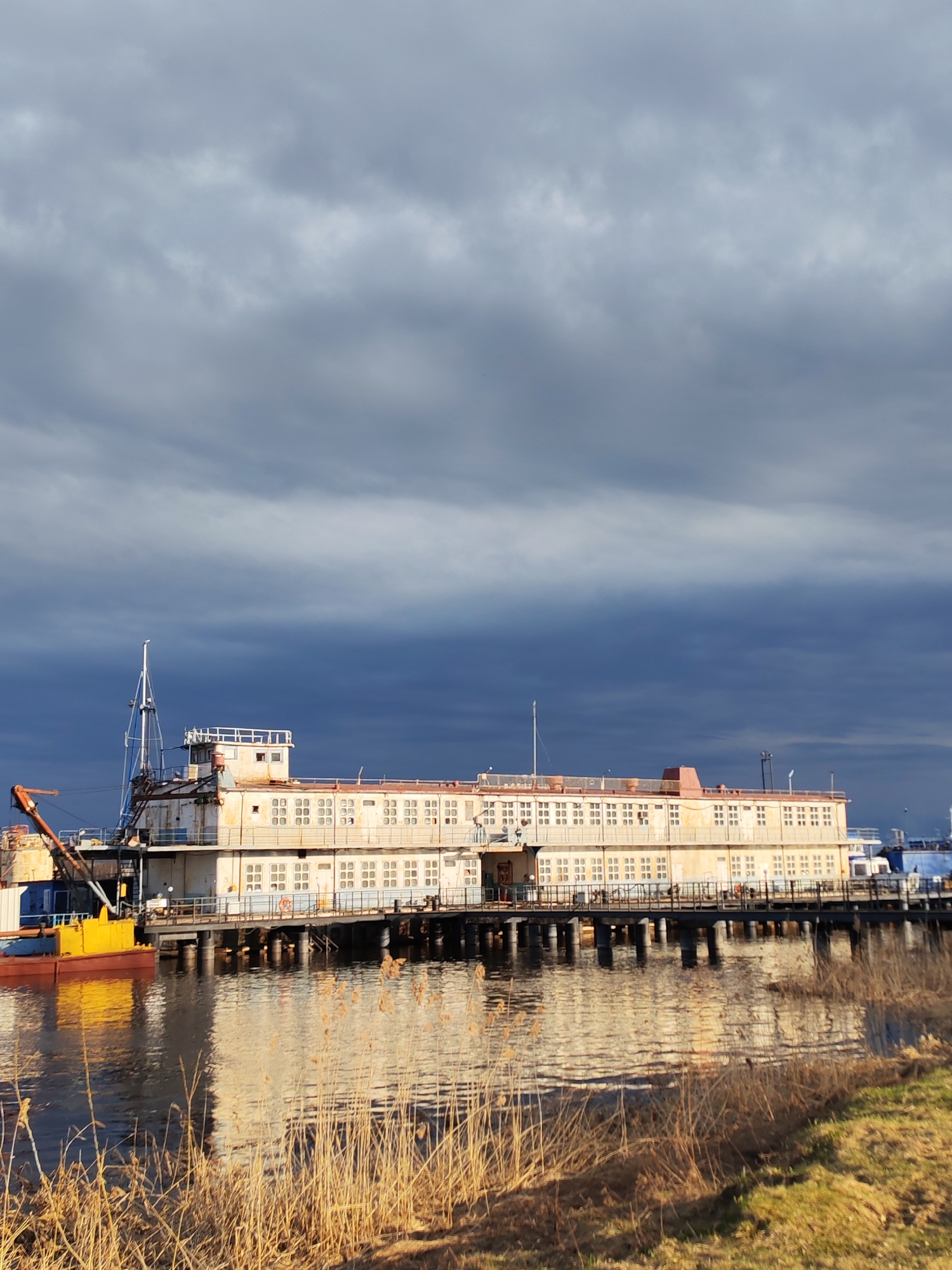 The image depicts a long, dilapidated, two-story building, likely a former river station or pier, situated on a body of water. The building is white with numerous windows and appears to be in a state of disrepair. A pier or walkway extends from the building over the water. A yellow and blue vessel with a crane is docked alongside the building. The water reflects the building and the sky. The foreground features tall, dry grass. The sky is dramatic with dark, heavy clouds. The location is Rybinsk, Russia.
