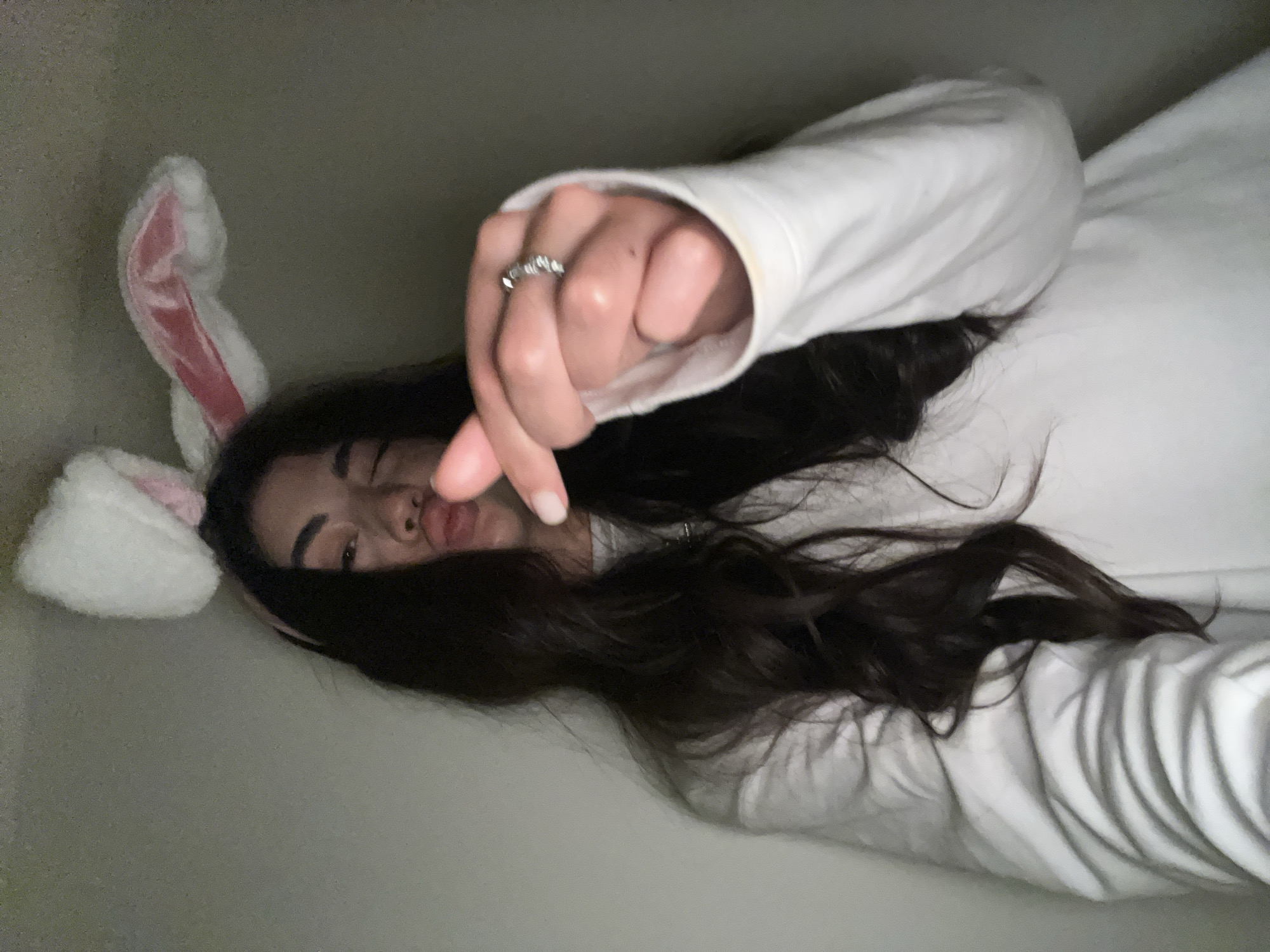 A young woman with dark hair, wearing a white sweatshirt and bunny ears, poses for a selfie in a dimly lit room. She is making a kissing face and holding her thumb and index finger together in front of her. A ring is visible on her left ring finger. The background is a plain, light-colored wall. There is no indication of the city of Chotěšov, Czechia.