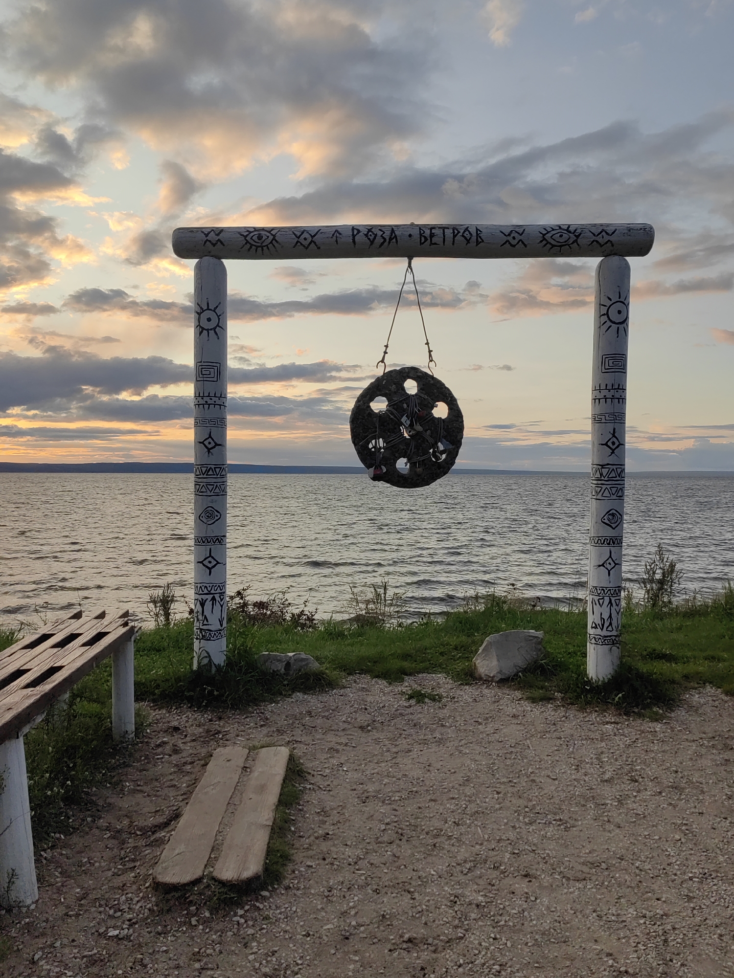 The image depicts an outdoor scene at sunset near a body of water, likely a lake or large river. In the foreground, there is a wooden bench and a dirt path with two wooden planks placed on it. The central focus is a wooden archway decorated with white paint and black symbols, resembling tribal art. Hanging from the archway is a circular object with multiple openings, possibly a decorative chime or a place for hanging locks. This archway is positioned overlooking the water, which is calm with visible ripples. The sky is filled with dramatic clouds in shades of orange, yellow, and gray, indicative of a sunset. Green grass and a few rocks are visible around the base of the archway. The overall atmosphere is serene and natural.
While the prompt specifies "city of Leninsky, Russia," there is no visual information within the image to confirm this specific location. The scene's details suggest a natural landscape that could be found in various regions.