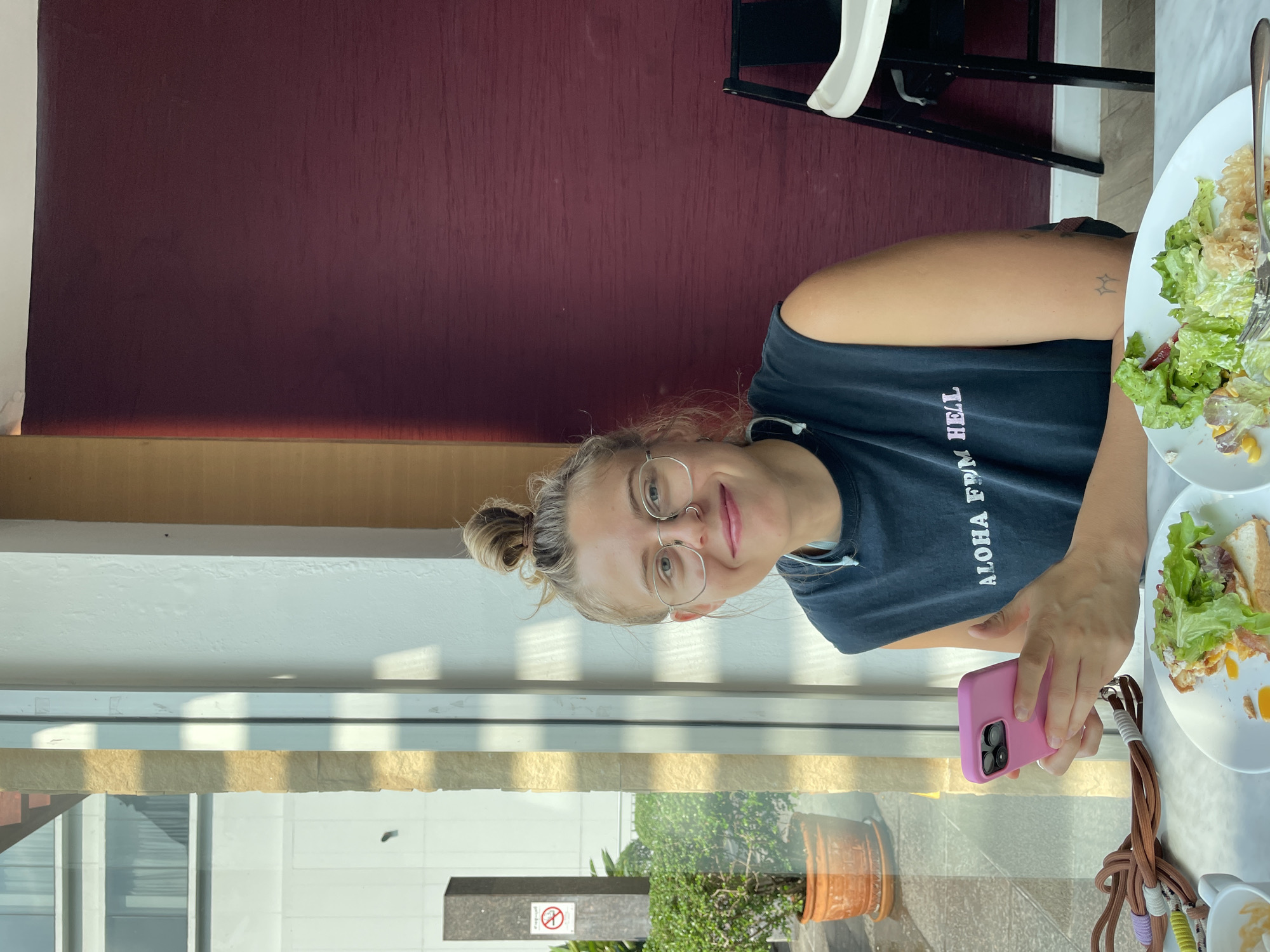 A young woman with light brown hair tied in a bun, wearing round eyeglasses and a dark t-shirt with "ALOHA FROM HELL" printed in white, smiles softly at the camera. She is seated at a table in what appears to be a cafe or restaurant in Bangkok, Thailand, indicated by the visible architecture and tropical plant outside.
Her right arm, adorned with a small, star-like tattoo, holds a pink smartphone. On the table to her right are two white plates, one holding salad leaves and a fork, the other with remnants of food, possibly from a sandwich. The table surface is light-colored, likely marble or granite.
Behind her, a dark maroon wall provides a warm backdrop. A black and white high chair is partially visible in the upper right corner. Below the table, a large window reveals an outdoor scene: a lush green potted plant, a concrete pillar with a "No Smoking" symbol, and modern building structures under bright daylight, suggesting a sunny day. The overall atmosphere is relaxed, capturing a meal or break time.