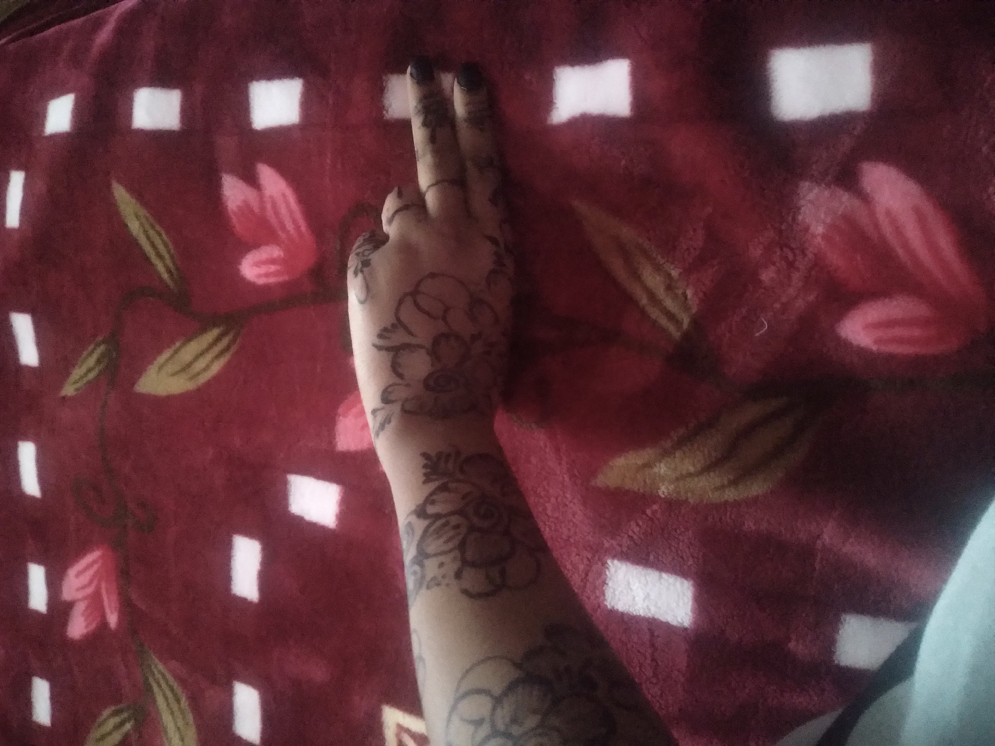 A close-up shot depicts a person's arm and hand adorned with henna tattoos, resting against a patterned burgundy blanket. The henna designs feature floral motifs. Two fingers are extended upwards. The person's nails are painted a dark color. The blanket exhibits a floral and geometric pattern with red, white, and gold colors. The location is unknown.