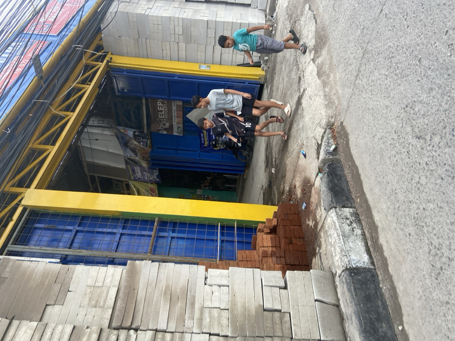 The scene is in front of a building with a yellow and blue striped awning. The building has brick walls with a yellow painted column on the left. There are a stack of red bricks at the base of the column. There are three boys in the scene. Two are walking away from the camera, one is wearing a soccer jersey and shorts and the other is wearing a blue t-shirt and black shorts. The third boy is walking towards the camera and is wearing a green t-shirt and black shorts. The boys are on the sidewalk. There is a motorcycle parked in the background. The scene is located in the city of Bojongsoang, Indonesia, at coordinates -6.972 latitude and 107.636 longitude.