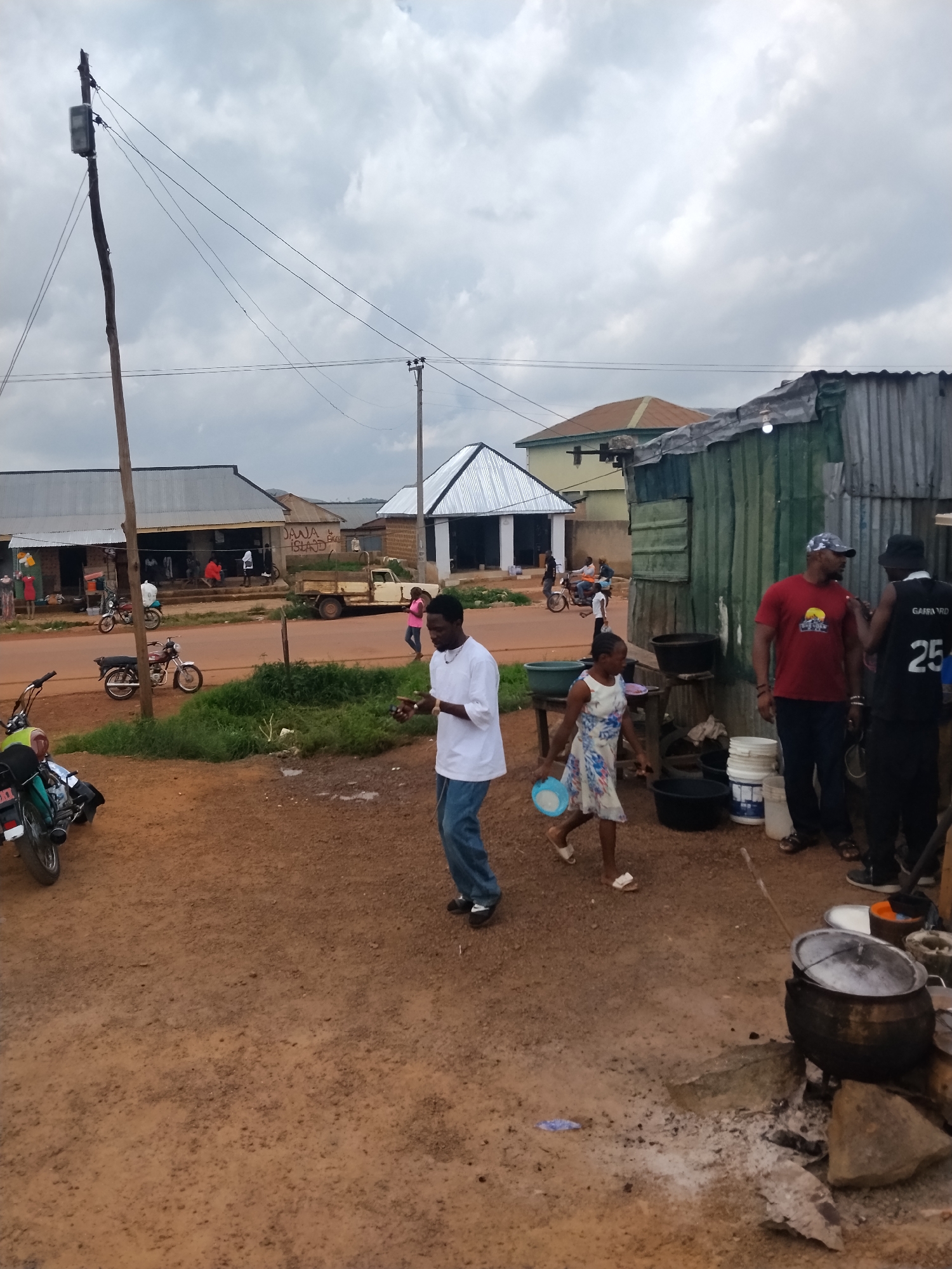 This is a street scene in Durumi, Nigeria. A group of people are gathered in front of small shops and makeshift structures. A man in a white shirt and blue jeans is walking away from the camera, holding a light blue bowl. A young girl in a floral dress is walking towards the camera, also holding a light blue bowl. A man in a red t-shirt and a man in a black vest with the number 25 are talking near a metal structure. Several large pots are arranged on a cooking fire in the foreground, with one pot containing water. Motorcycles are parked along the side of the road, and a light-colored pickup truck is visible in the background. The ground is dirt and gravel, and the sky is overcast. The overall impression is a bustling, everyday scene in a Nigerian community.