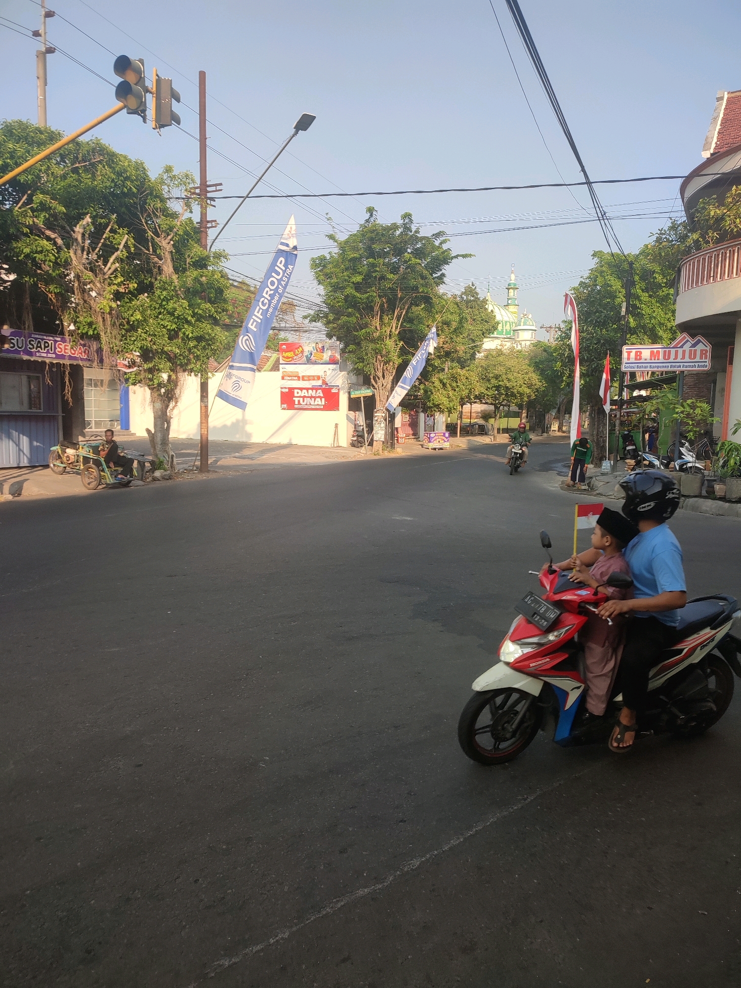In Nganjuk, Indonesia, on a paved street bathed in the soft glow of morning light, a scene unfolds at a street intersection. Two individuals, a man and a child, ride a red and white motorcycle in the foreground. The man, wearing a blue shirt and black pants, is at the helm, while the child, dressed in a pink outfit and a black cap, sits behind him. Both wear helmets, with the man sporting a black one.
To the left, a man in a black shirt and pants rides a three-wheeled motorcycle, stopped near a building with a blue facade. A white banner with blue text, "FIFGROUP," is visible. Across the street, a mosque with green domes and a tall minaret stands under a clear blue sky, its silhouette a distant yet prominent feature. Several trees with lush green foliage line the street, their branches casting shadows on the asphalt.
Several businesses are visible. One sign reads "SU SAPI SESAR," and another, a red banner, displays "DANA TUNAI." A sign with "TB. MUJJUR" and "Solusi Bahan Bangunan Untuk Rumah Anda" is also present, suggesting a building materials store. Traffic lights hang overhead, with yellow and red lights illuminated, indicating a controlled intersection. Electric wires crisscross the sky, a common urban sight. The overall impression is a typical morning commute in a bustling Indonesian city.