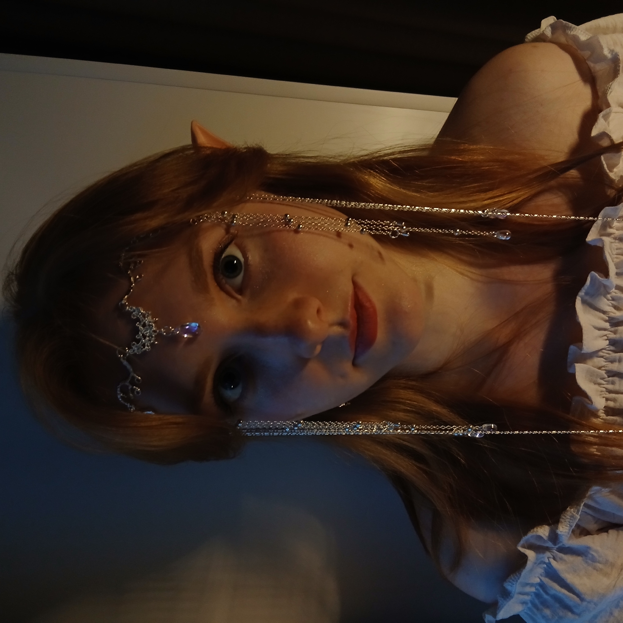 The image, rotated 90 degrees clockwise, captures a young person, likely female, with long reddish-brown hair and fair skin. They are looking directly at the camera with wide, dark eyes and a soft, subtle smile. A notable feature is their pointed ears, suggesting an elf or fantasy character.
A delicate silver chain headpiece adorns their head, featuring intricate designs, small clear beads, and a prominent purple jewel near their left temple. The chains drape gracefully across their forehead and down the sides of their hair. A ruffled white garment, possibly a blouse or dress, is visible on their right shoulder and chest.
The lighting is dim, casting dramatic shadows and highlighting the left side of their face and hair. The background is largely dark and indistinct, with a lighter, possibly off-white, wall section visible behind their head and shoulder. The person appears to be posing for a photograph. The overall impression is one of ethereal beauty, perhaps for a fantasy-themed event or portrait. No text is visible in the image, and specific location or weather cues are absent.
