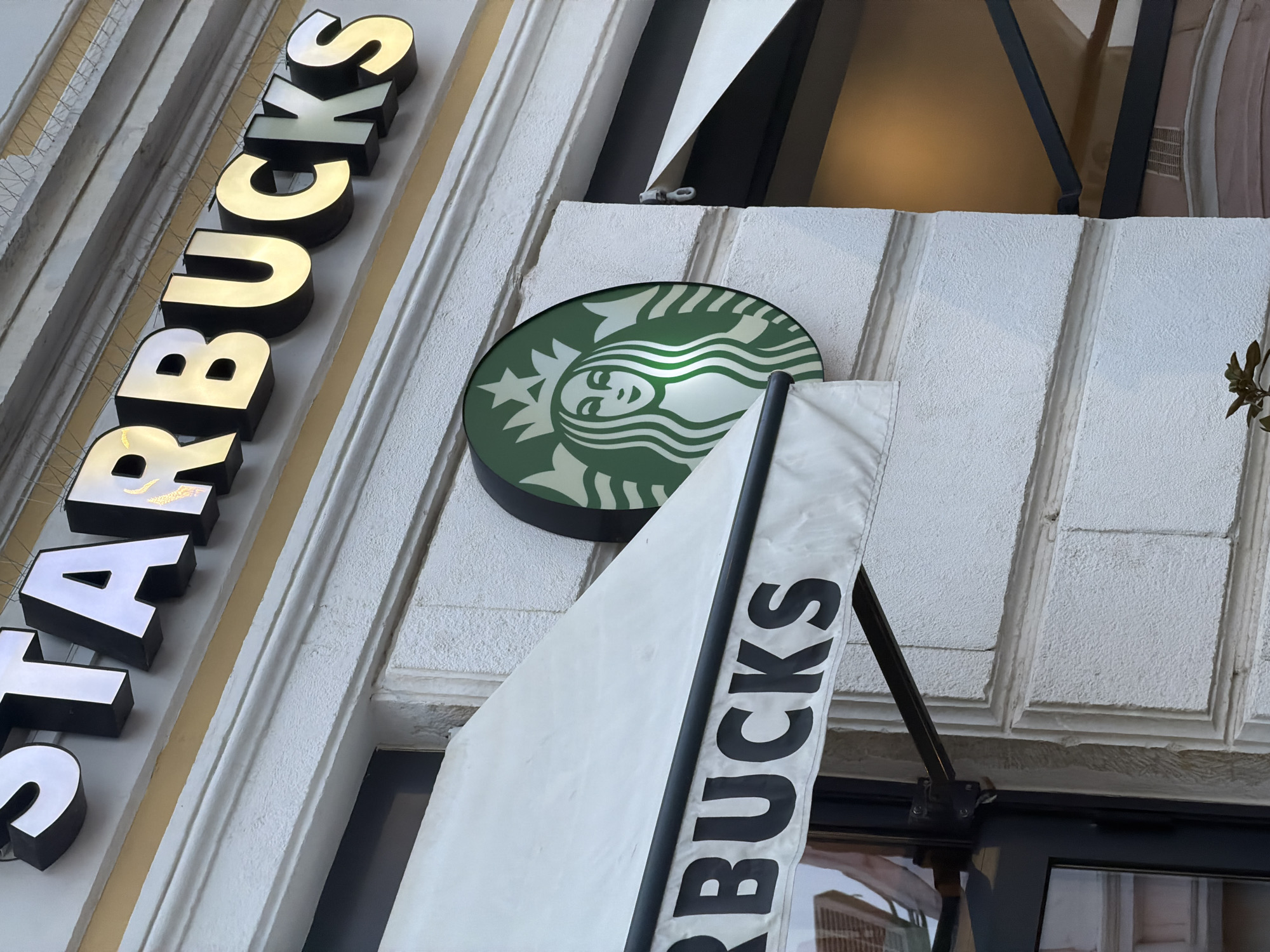 The image shows a Starbucks store facade in Varna, Bulgaria. The building is white with decorative moldings. The Starbucks logo, a green circle with a stylized white siren, is prominently displayed above the entrance. The word "STARBUCKS" is vertically written in illuminated letters on the side of the building. There is also a white fabric banner displaying the word "BUCKS". The architectural style of the building appears to be influenced by European design.