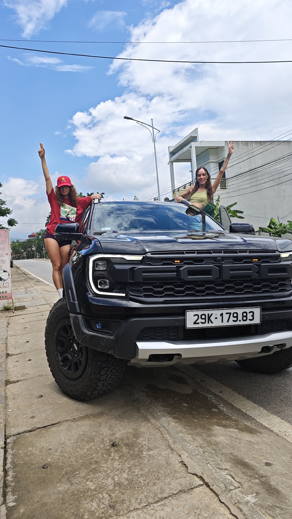 Two women are posing with a black Ford pickup truck in Trung Tam Ward, Vietnam. One woman, wearing a red shirt and hat, stands beside the truck, leaning against the driver's side. The second woman stands in the truck's cab, with her upper body protruding through the open roof. Both women are smiling and raising their arms. The truck, identified by its license plate "29K-179.83," is parked on a paved surface near a roadside. In the background, there are buildings, power lines, and trees under a partly cloudy sky. A sign with Vietnamese script is visible in the background.