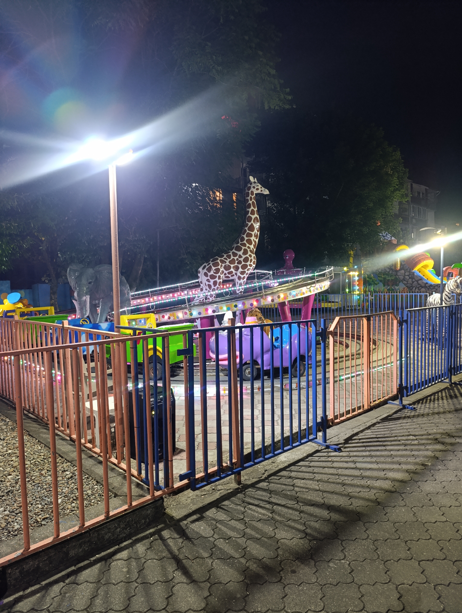 This is a night photograph of a children's amusement ride in Batumi, Georgia. The ride features colorful animal figures, including a giraffe, an elephant, and a zebra, and appears to be a spinning or rotating carousel-style attraction. The ride is enclosed by a fence with orange and blue vertical bars. The ground is paved with interlocking stones, and there is gravel to the left of the fence. Trees are visible in the background, and the scene is illuminated by bright lights, casting long shadows. No people are visible in the photograph.