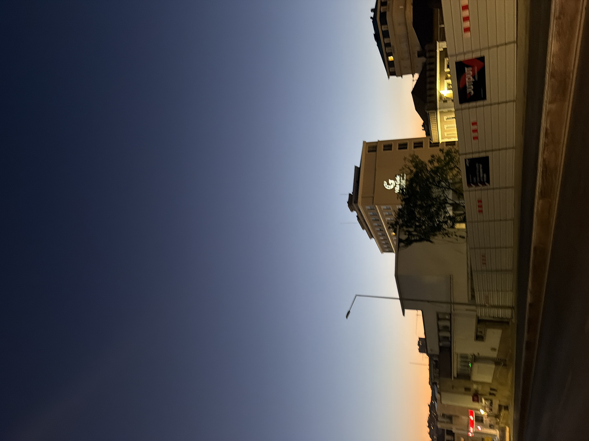 The image depicts a twilight scene in Cascais, Portugal, with a deep blue sky fading into a warm orange hue near the horizon. In the mid-ground, several buildings are visible. A prominent, light-colored building with multiple stories features a lit logo that reads "Villa Gatte." To its right, another building with a darker facade and illuminated windows is visible. In the foreground, a construction barrier runs across the scene, displaying advertisements, one of which says "OUTPUT." A street lamp is present on the left side of the image. The overall environment suggests an urban setting at dusk, with streetlights and building lights beginning to illuminate the scene. There are no people clearly visible in the image.