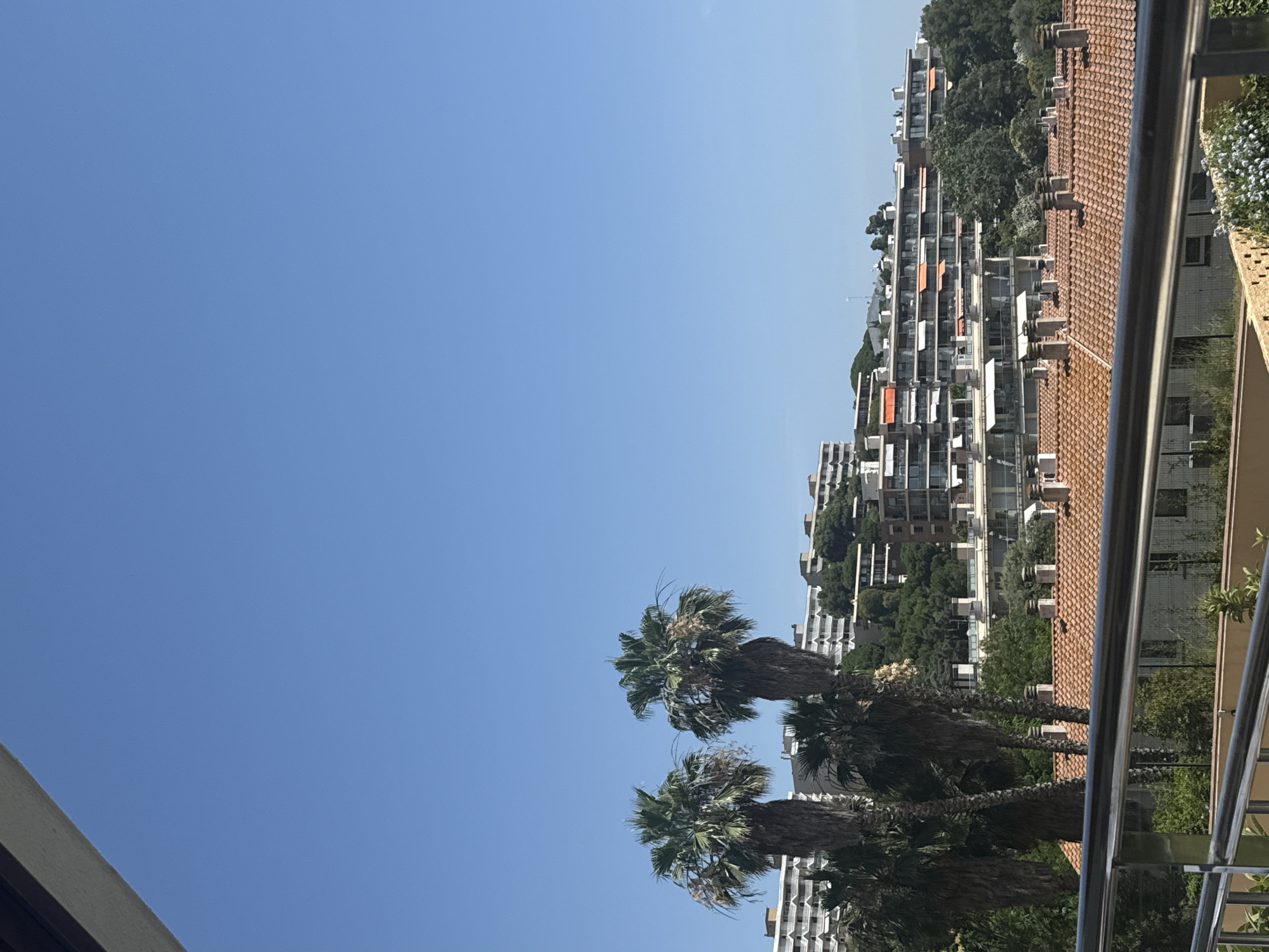 This media file captures a view from a balcony in Sant Feliu de Guíxols, Spain. The scene is dominated by a clear blue sky, indicating it is daytime with fair weather. In the foreground, three palm trees stand tall, their fronds rustling gently, suggesting a pleasant, possibly warm, atmosphere.
Behind the palm trees, a hillside is densely packed with buildings, characteristic of a Mediterranean coastal town. The architecture appears to be a mix of modern apartment complexes with white facades, balconies, and some orange awnings, interspersed with older structures featuring red-tiled roofs. The buildings ascend the hill, creating a layered effect. Lush green trees are visible between and behind the buildings, adding a natural element to the urban landscape.
A metal railing runs along the bottom right of the frame, implying the perspective is from a raised vantage point, such as a balcony or terrace. The overall impression is one of a sunny, relaxed day in a scenic coastal town. There are no people or specific activities depicted; the focus is on the landscape and architecture. No visible text is present in the image.