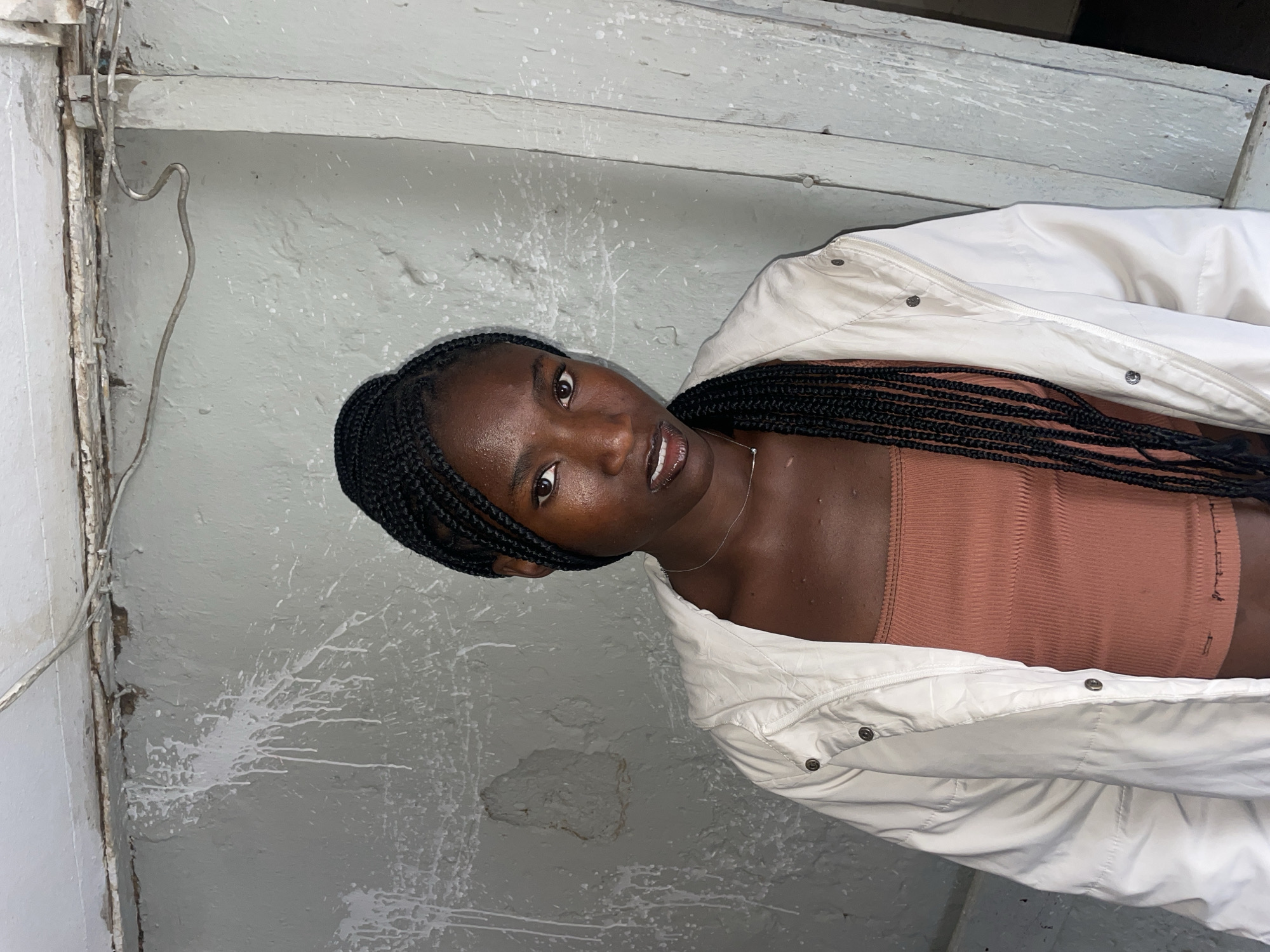 A portrait photograph, likely taken indoors or in a sheltered outdoor space in Jos, Nigeria, features a young Black individual with long, dark box braids. The subject is looking directly at the camera with a calm and direct expression. They are wearing a form-fitting, ribbed bandeau or tube top in a warm brown/tan color, layered under a white, unzipped jacket. A thin silver necklace is visible around their neck. Their skin exhibits a noticeable sheen, possibly from sweat, oil, or a hydrating product.
The background consists of a textured, light grey wall, showing signs of wear and tear including patches of peeling paint and numerous white splatters, giving it a weathered appearance. Along the left edge of the frame, a thin, bent metallic wire runs vertically, fastened to the wall. The lighting in the image is even and soft, highlighting the subject's features and the rough texture of the wall. No discernible text is visible within the frame. The overall atmosphere is candid, focusing on the individual against a utilitarian and somewhat aged backdrop.