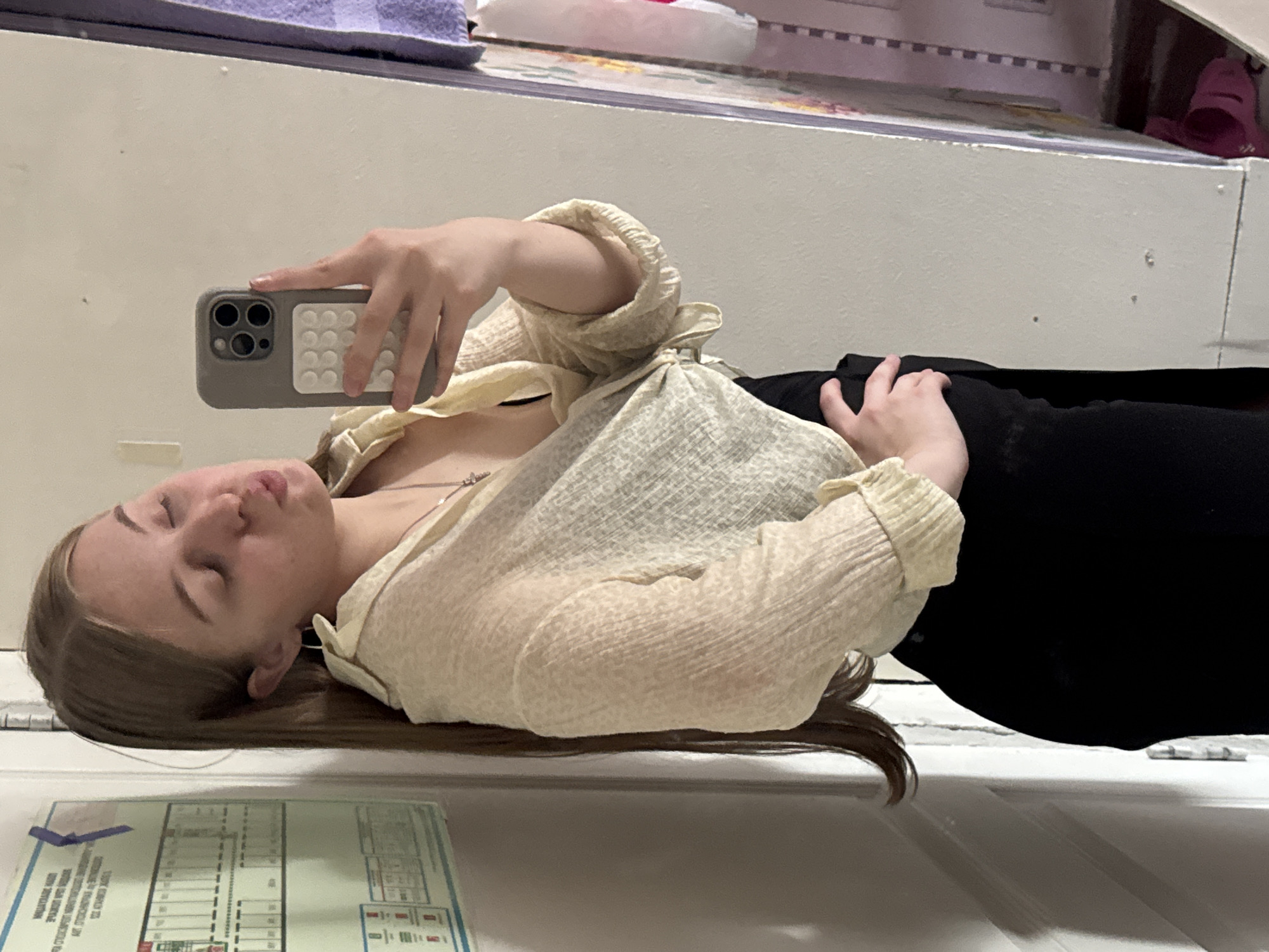 A young woman with long, light brown hair is taking a mirror selfie in what appears to be a dimly lit room. She is wearing a sheer, light yellow, button-down shirt tied at the waist, revealing a black undergarment, and black pants. Her eyes are closed, and her lips are pursed as if she is blowing a kiss. She has a silver necklace with a small pendant. In her right hand, she is holding a smartphone with a gray case. To the left of the woman, a white door with a poster attached to it is visible. The poster contains a grid-like structure with text in Russian, possibly an emergency exit plan or evacuation map, common in public buildings or shared living spaces. To the right of the woman, a portion of a closet or shelf unit is visible with colorful items and a pink shoe on the floor. The overall lighting suggests an indoor setting, likely an apartment or residential building. The presence of the evacuation map is a common feature in buildings in Perm, Russia, and other parts of the country.