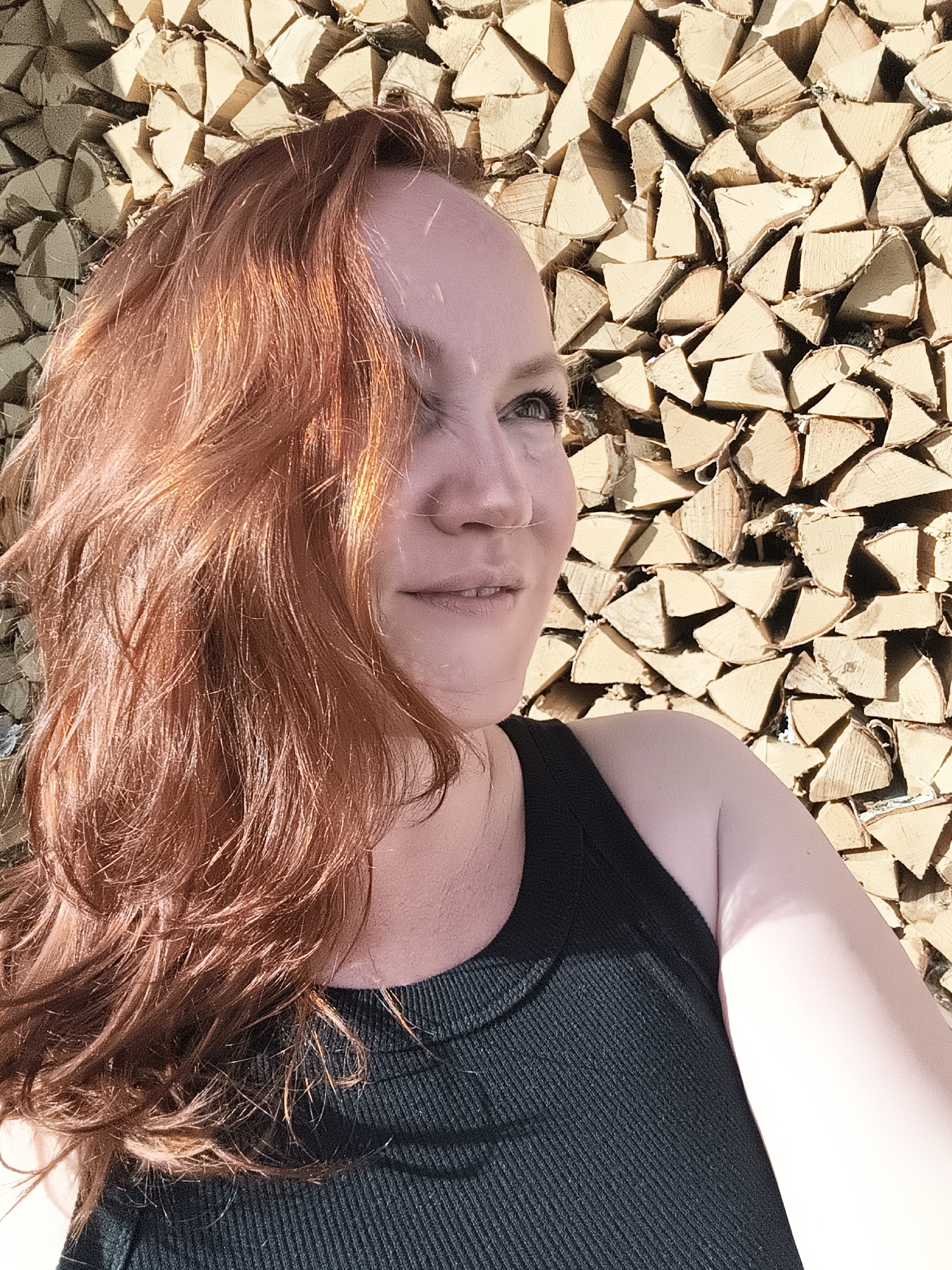 A woman with long, wavy, ginger hair is pictured in a close-up, outdoor shot. She is looking off to the right, with a slight smile on her face. Her left eye is visible, and her skin appears fair with freckles. She is wearing a dark, ribbed tank top. Behind her, a large, neatly stacked pile of firewood serves as the backdrop. The wood is cut into logs of various sizes, and the sunlight casts shadows across the uneven surfaces, suggesting it is daytime. The overall lighting is bright and warm, indicating sunny weather. The context places this scene in Chaikovsky, Russia. No text is visible in the image.