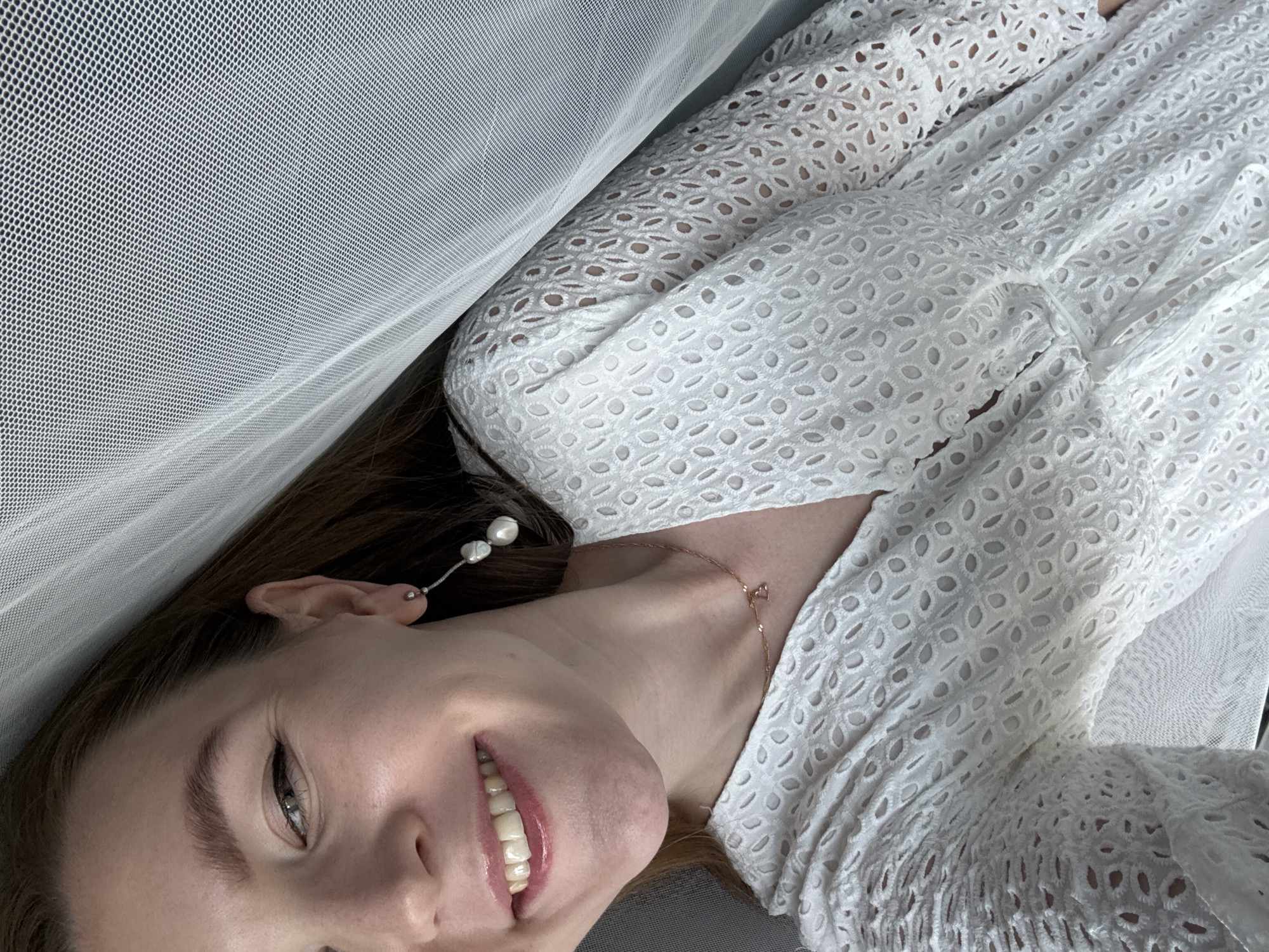 The image shows a close-up of a woman's face and upper body. She is smiling and looking towards the camera. She has long brown hair, and is wearing a white dress with an eyelet pattern. She is also wearing a necklace with a small pendant, and a long earring with two pearls. The background is out of focus, but appears to be a white, sheer fabric, possibly a curtain or veil, and a gray surface. The lighting suggests an indoor setting, possibly near a window. There is no information in the image that would allow for the determination of its location in Izhevsk, Russia.