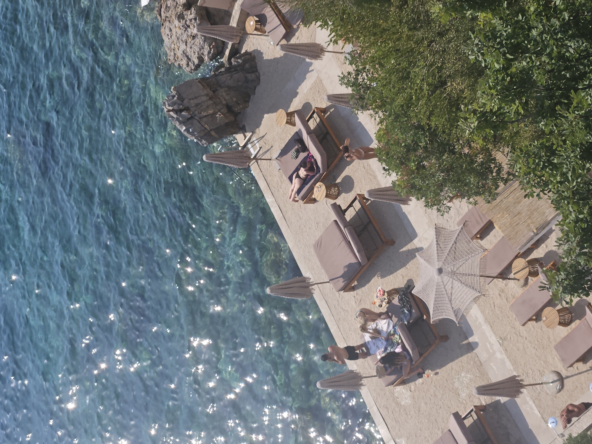 This high-angle aerial shot captures a sun-drenched beach area adjacent to the clear, turquoise waters of the Adriatic Sea, likely in Budva, Montenegro. The scene is populated with several lounge chairs, many with patrons relaxing.
In the foreground, a man in black shorts stands near the water's edge, looking away from the camera. Beside him, a group of individuals are seated on a lounge chair covered with a colorful towel, engaged in conversation. Further down the beach, a woman in a bikini reclines on a double lounge chair. Another woman, also in a bikini, stands near this lounge, appearing to interact with someone off-camera.
Scattered throughout the beach are several beige beach umbrellas, some closed and some open. Small wooden side tables are positioned next to some of the lounge chairs, with some holding drinks or small items. In the background, rocky outcrops meet the sea. Lush green trees frame the right side of the image, providing a contrast to the sandy beach and blue water. The water itself is filled with shimmering reflections from the sun. The overall impression is one of leisure and relaxation in a beautiful coastal setting.