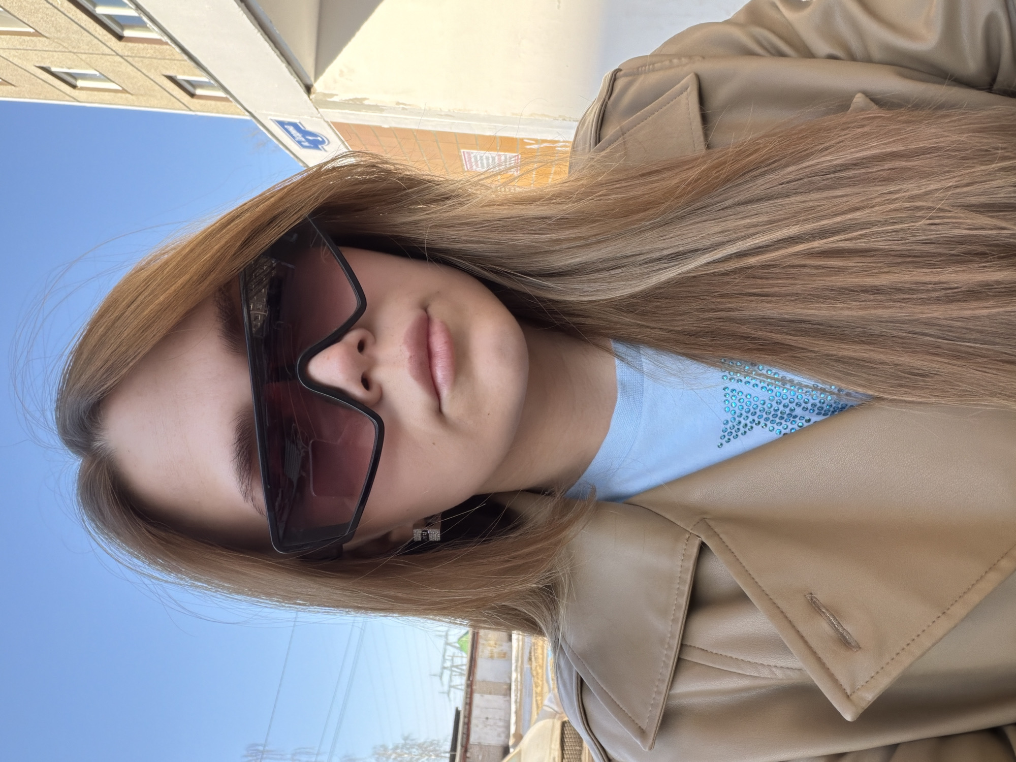 A woman wearing sunglasses and a tan leather jacket is posing for a selfie. She has long, blonde hair and is wearing a light blue t-shirt with a star pattern. In the background, there is a building with windows, possibly an apartment building, and a blue sky. Based on the partial view of the street sign, the location could be identified as Chaikovsky, Russia. Other details in the background include a fence, buildings, and what appear to be power lines. The photo is taken at an angle, and the environment appears to be outdoors during the daytime.