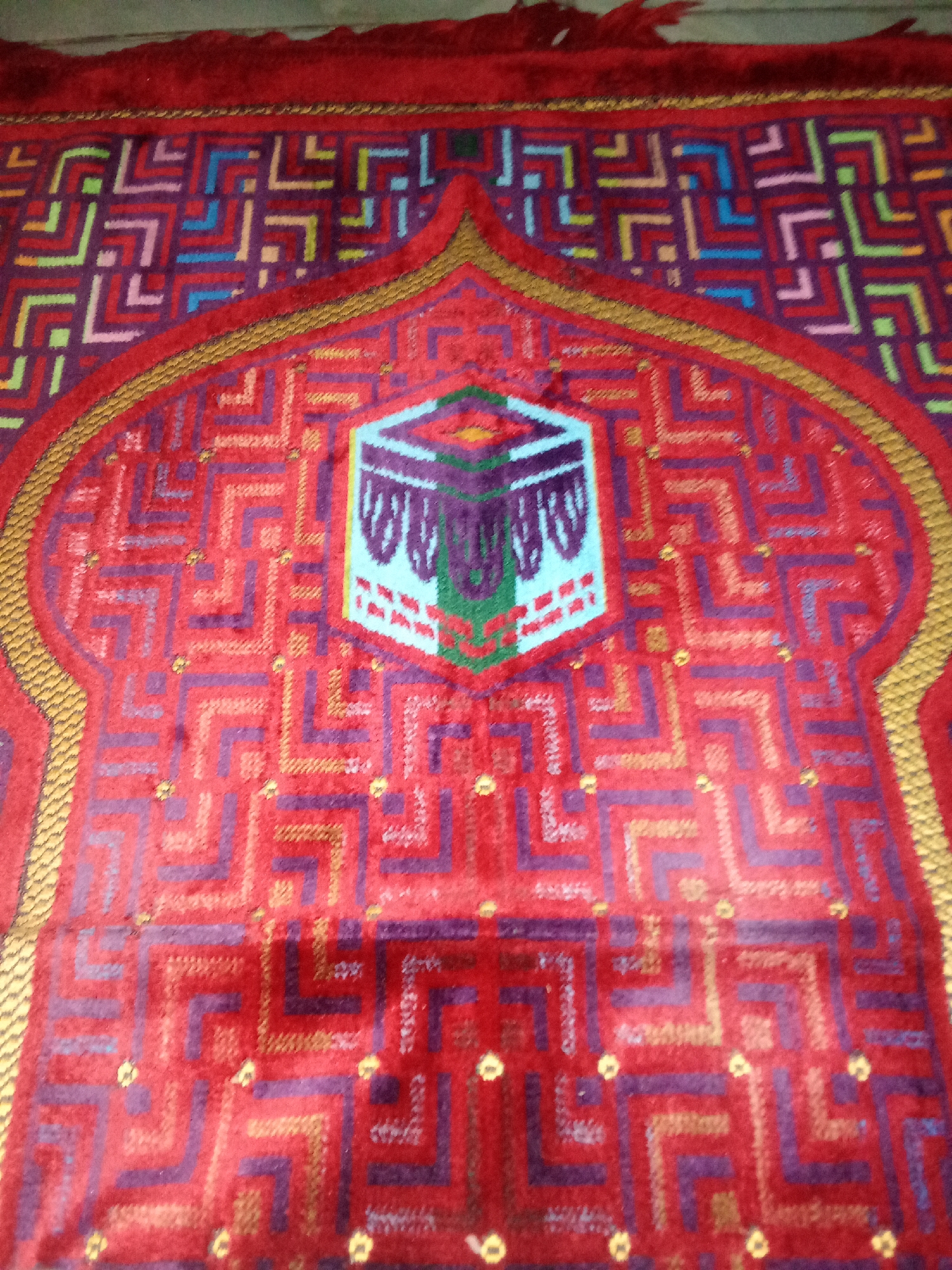 This is a close-up, slightly blurry, top-down shot of a prayer rug. The rug is predominantly red with an intricate geometric pattern in purple, blue, and some lighter colors. In the center of the rug, there is a stylized representation of the Kaaba, a cube-shaped building in Mecca, Islam's holiest city. The Kaaba is depicted with a light blue facade and purple draped fabric, suggesting it is covered with the kiswah. The rug has a tasseled red edge visible at the top and a gold embroidered border framing the central arch-like design. The image was taken in Cempaka, Indonesia.