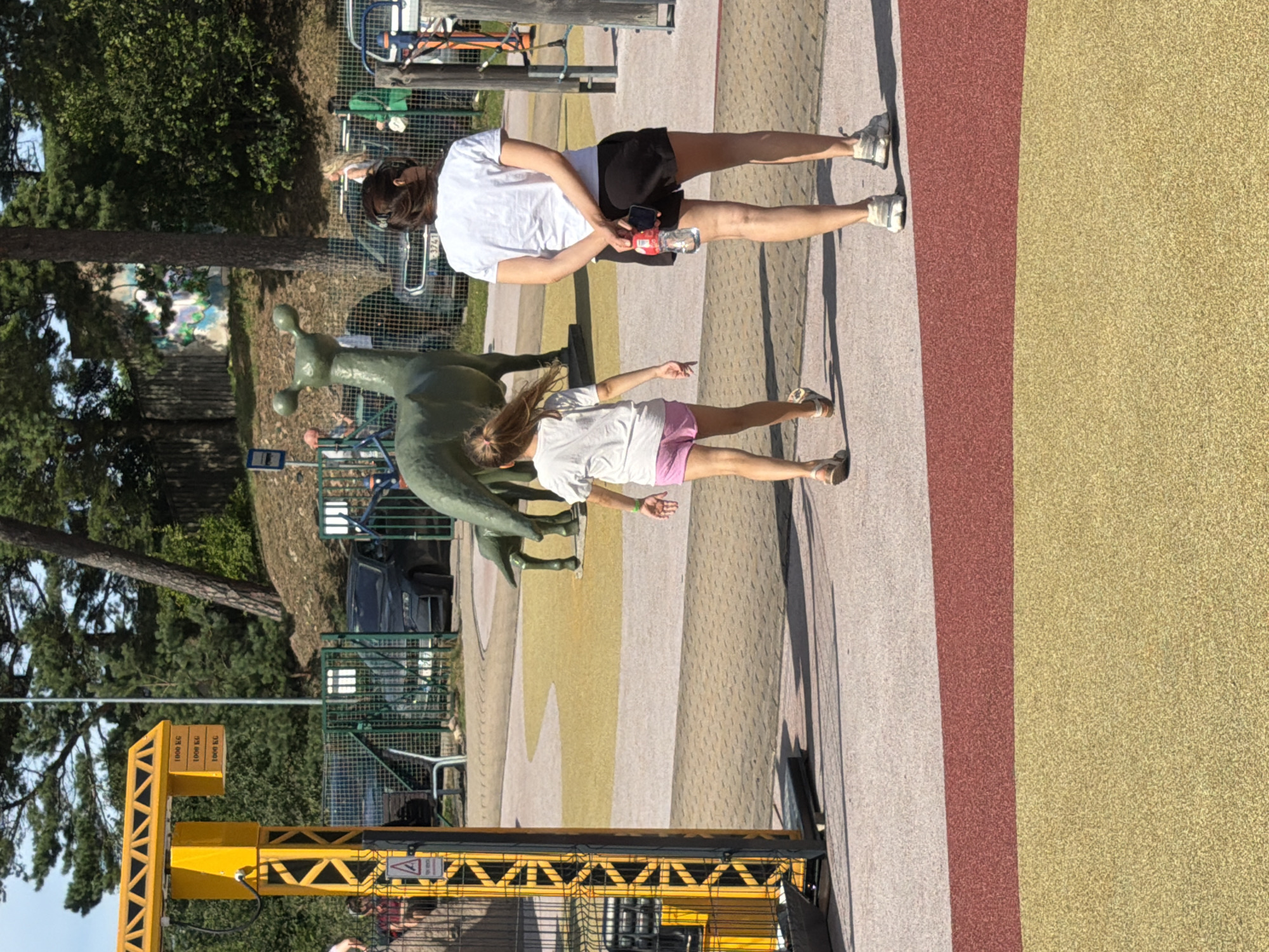 The image shows an outdoor scene with a yellow construction crane-like structure on the left. The ground is paved with interlocking stones, and there are areas of colored rubber surfacing, typical of a playground. In the background, there is a metal fence, trees, and some vehicles.
In the foreground, two people are walking away from the camera. On the right is a woman wearing a white t-shirt and black shorts, holding a bottle of water and a phone. To her left is a young girl wearing a white t-shirt and pink shorts. They are walking on the paved surface, with the girl slightly ahead of the woman.
Behind them, in the middle ground, is a green sculpture of a long-necked creature, possibly a dinosaur or mythical animal, with a smaller, similar creature behind it.
The overall scene appears to be a public space, possibly a park or recreational area. This specific location is a playground in the Ventspils Adventure Park in Ventspils, Latvia. The yellow structure is part of an exhibit related to the park's maritime and industrial history.