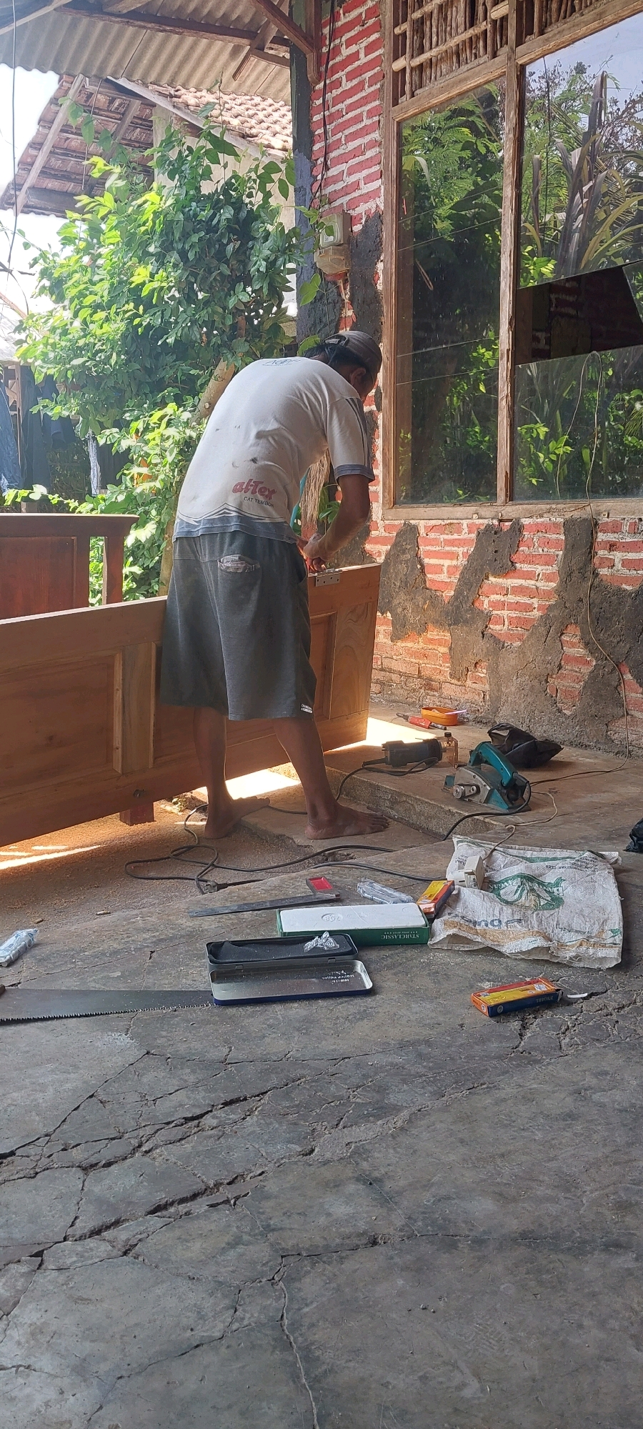 In the city of Keling, Indonesia, a person is working on a wooden structure. They are wearing a white t-shirt and gray shorts and appear to be attaching a hinge to a wooden door or panel. Tools such as a hand saw, a power planer, and other unspecified items are scattered on the concrete floor, which is cracked. Behind the person, a red brick wall is visible, with part of a wooden-framed window that reflects the surrounding greenery. The scene takes place outdoors, under a roof with corrugated sheeting, with lush foliage evident in the background.