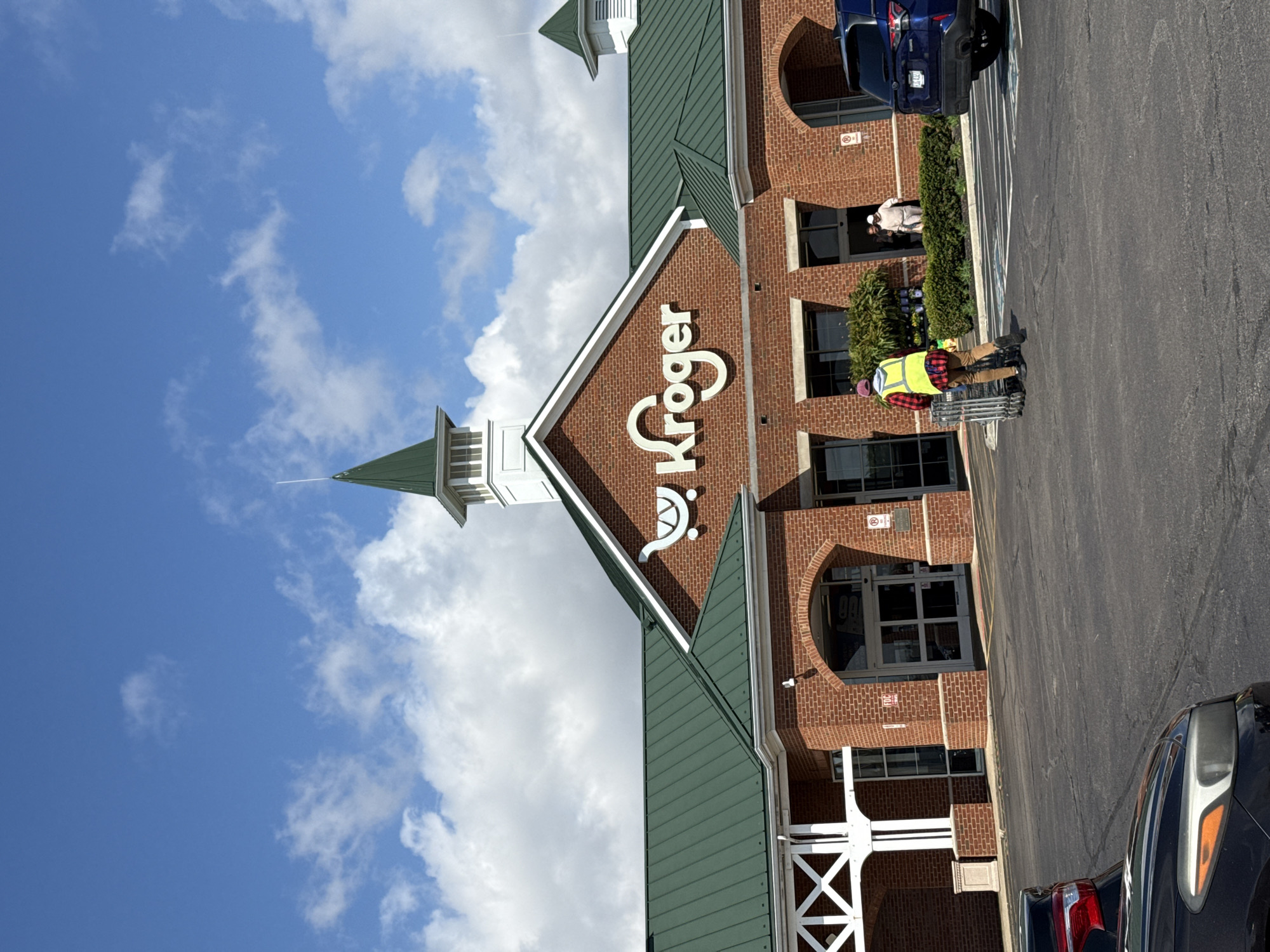 The image shows the exterior of a Kroger grocery store in Columbus, Ohio, USA. The building is made of red brick with a green roof and a distinctive steeple. The Kroger logo is prominently displayed. A man wearing a safety vest is pushing a shopping cart in the parking lot. A few cars are parked in the lot. The sky is blue with some clouds.