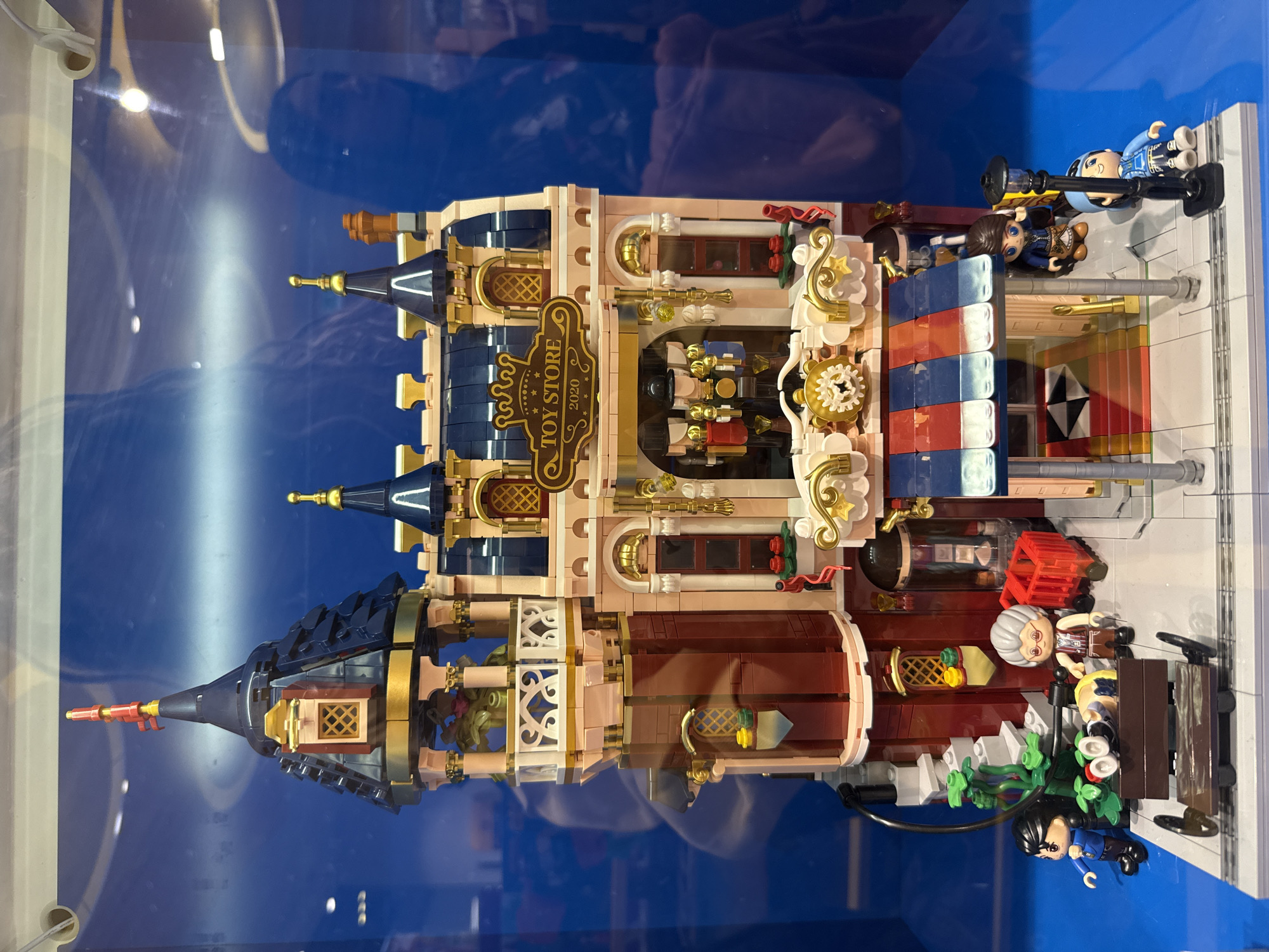 This image displays a detailed Lego model of a toy store. The building is multi-story, with a dark blue roof featuring spires and gold accents. The facade is a mix of pink and reddish-brown bricks, with ornate gold trim around windows and doorways. A prominent sign above the entrance reads "TOY STORE 2020". The ground level features a striped awning over the entrance. Several Lego figures are present: on the left, a figure in a blue jacket stands near a bench with another figure lying on it. An elderly figure with grey hair stands to the right of the bench, next to a small red cart. Two figures are positioned by the entrance, and another figure stands near a lamppost. The model is displayed on a grey base with a brick pattern. The background is a solid blue, suggesting a display case or backdrop. The image does not contain any information that would indicate the location in the city of Yaroslavl, Russia.