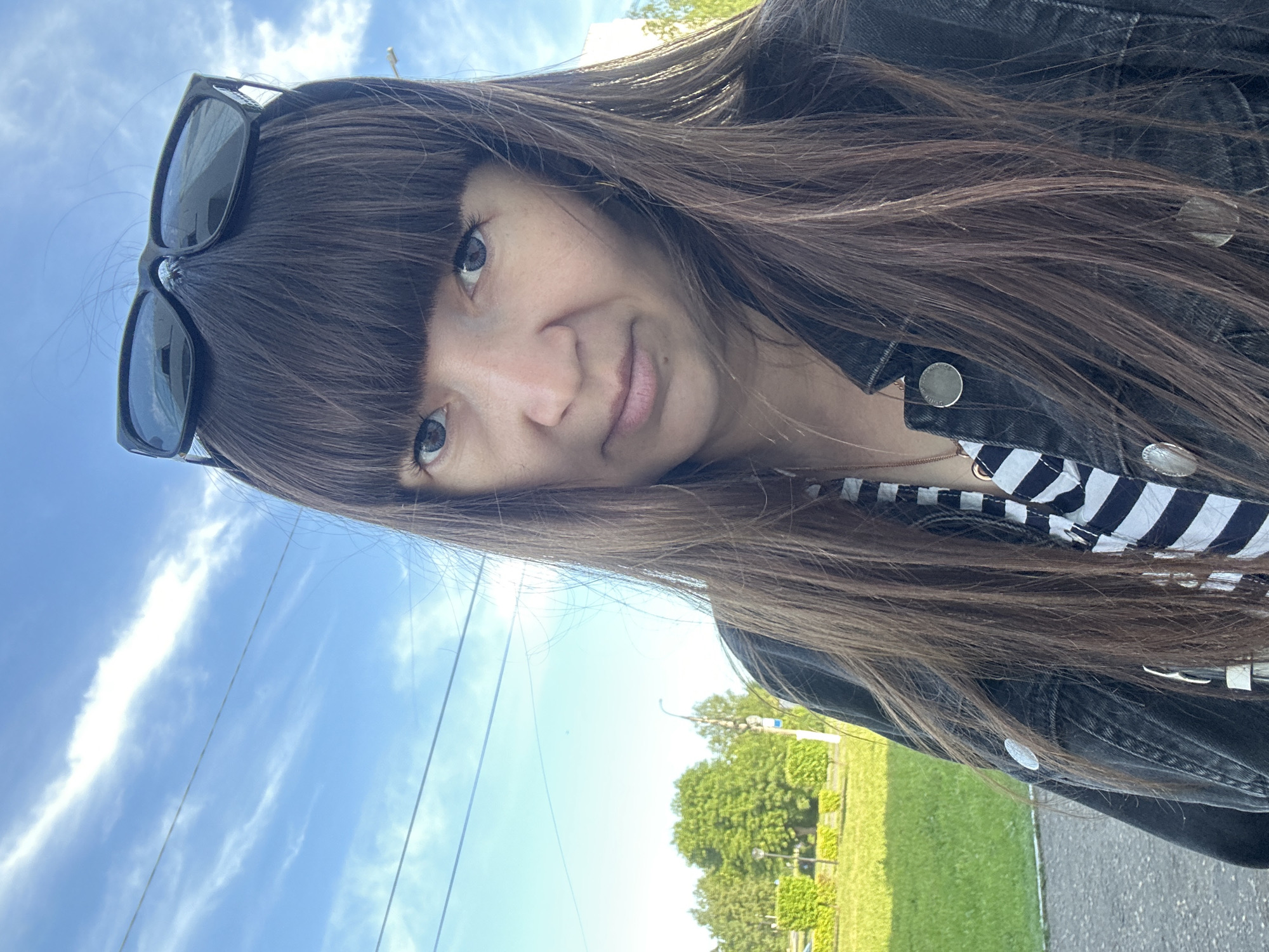 A young woman is featured in an outdoor selfie, taken in Chaikovsky, Russia. The image is oriented with the woman's head tilted significantly, appearing rotated 90 degrees counter-clockwise. She has long, straight dark brown hair with bangs that cover her forehead. Her light-colored eyes, possibly blue or grey, gaze directly at the camera, and her lips form a subtle, slight smile or smirk. Black-framed sunglasses are pushed up and rest on top of her head, nestled within her hair. She is dressed in a dark, likely black, denim jacket with several metallic silver buttons or snaps visible, worn over a top with black and white horizontal stripes.
The background shows a clear, bright blue sky with scattered wisps of white clouds, suggesting it is daytime with pleasant weather. Thin, dark lines, likely power lines or telephone wires, stretch across the sky. Green foliage from trees and grass fills the lower and right parts of the background (when viewed upright), indicating an outdoor setting such as a park or along a street. A dark paved surface, resembling a road or sidewalk, is also partially visible. The overall scene captures a relaxed, casual moment. There is no visible text in the image.