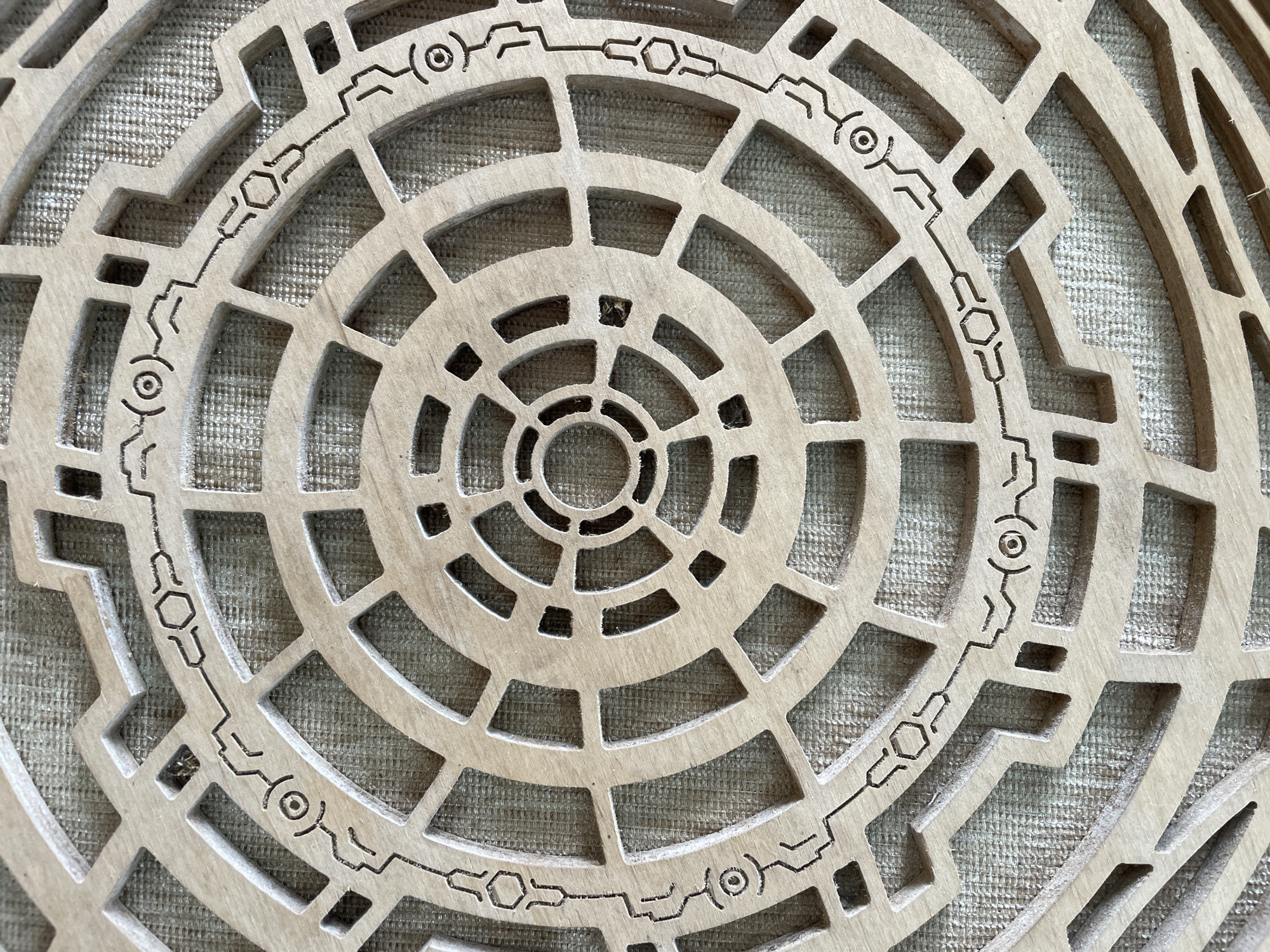 The image shows a close-up, overhead view of a circular wooden object with intricate geometric carvings. The object is placed on a textured fabric surface, possibly burlap or canvas, which appears to be a light brown or olive green color. The wooden object is light brown and features concentric circles and radial lines that create a segmented pattern. Some segments are filled in, while others are open, allowing the fabric to show through.
Along the outer edge of the circular object, a thin, dark line forms a decorative border. This border contains various abstract symbols, including what appear to be stylized eyes, hexagons, and connecting lines. These symbols are carved into the wood and filled with dark pigment, contrasting with the natural wood grain. The carving technique appears to be laser-cutting or a similar precise method. The texture of the fabric is visible through the open spaces in the wooden design. There are no people or animals visible in the image, and the scene is static. The lighting is even, highlighting the details of the carving and the texture of the materials. The image does not provide any information about the location of Bhagsu, India.