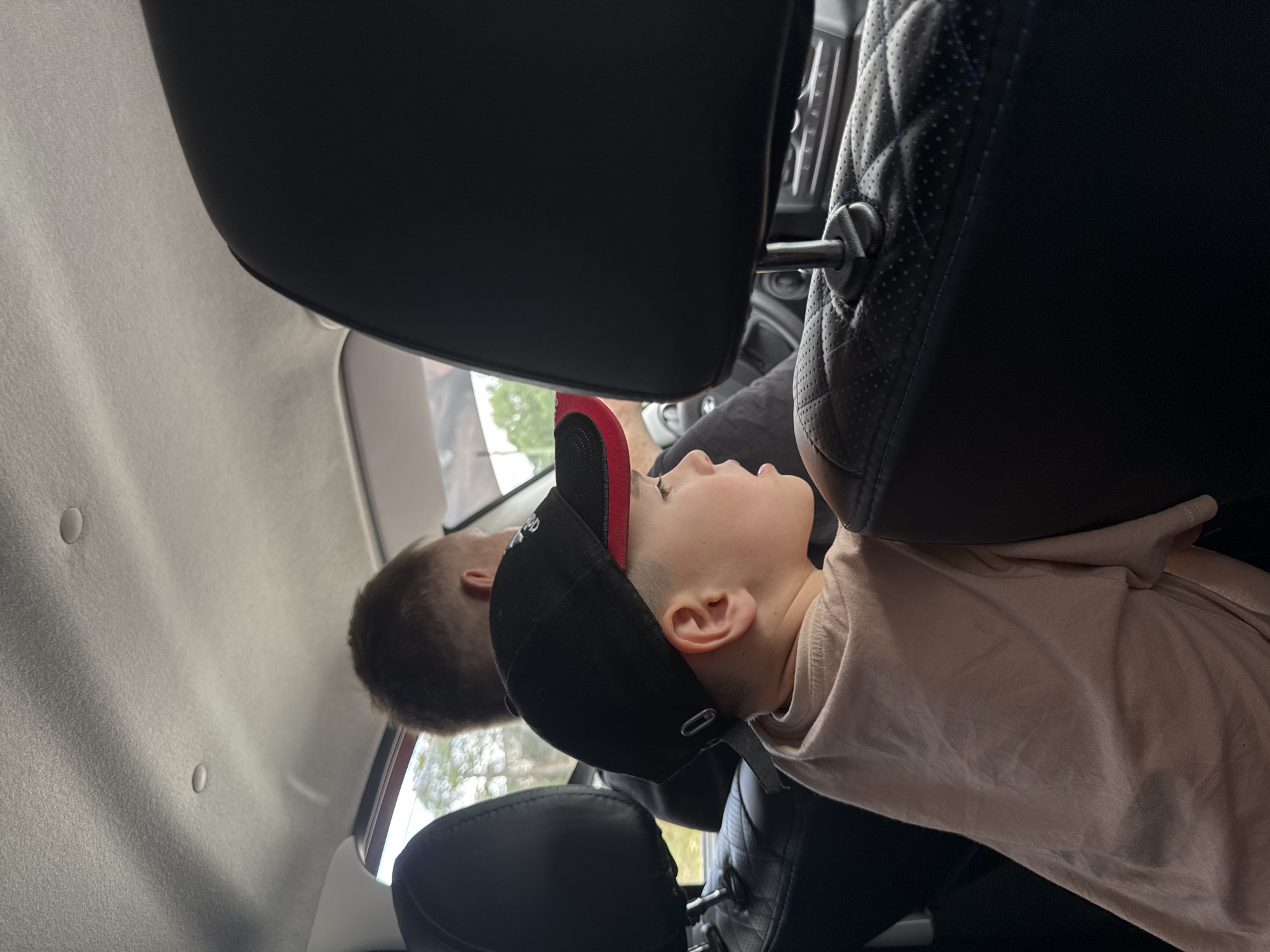 A medium shot shows a young boy in a black baseball cap with red trim and a light pink t-shirt sitting in the back seat of a car, looking to his right. His face is partially obscured by his cap and the car's headrest. Behind him, the back of a man's head and shoulders are visible. The interior of the car is dark, with black leather seats and a light-colored headliner. Through the window, trees and foliage are visible. The scene is set in Berezovaya Staritsa, Russia.