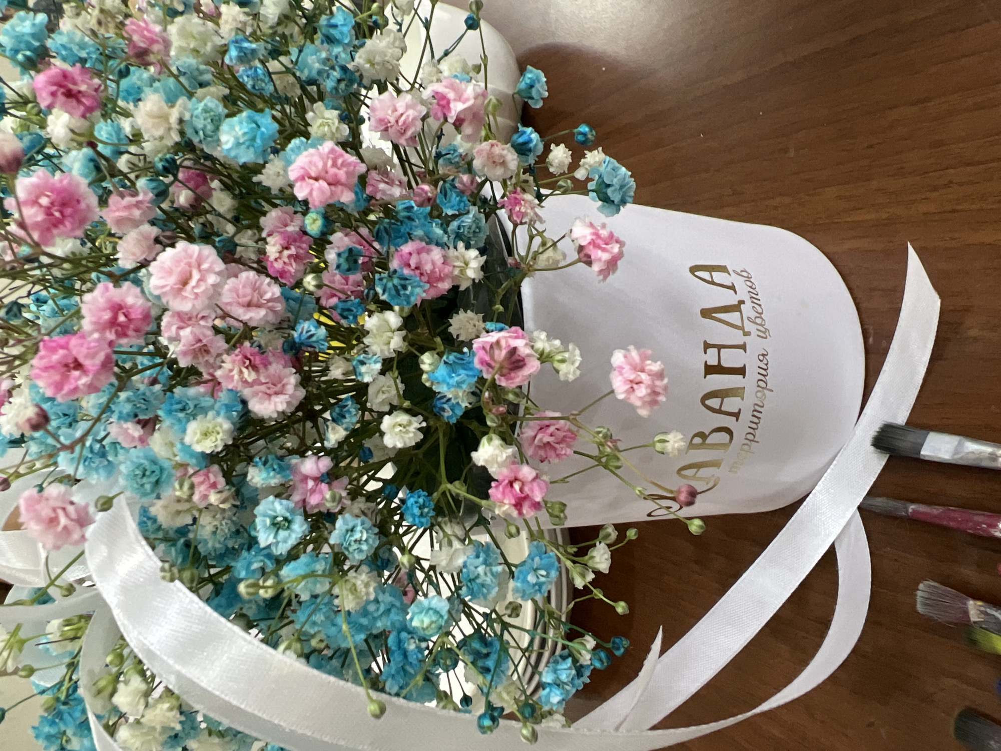 A floral arrangement featuring pink, blue, and white gypsophila flowers fills a white, cylindrical container. The container is decorated with text in gold lettering. A white satin ribbon is tied around the container. The scene appears to be set on a wooden surface. Paintbrushes lie next to the ribbon on the right side. The Cyrillic text on the container appears to say "Abada, территория цветов" ("Abada, territory of flowers"), which may refer to a flower shop of that name. There is a location of such a business in the city of Chaikovsky, Russia.