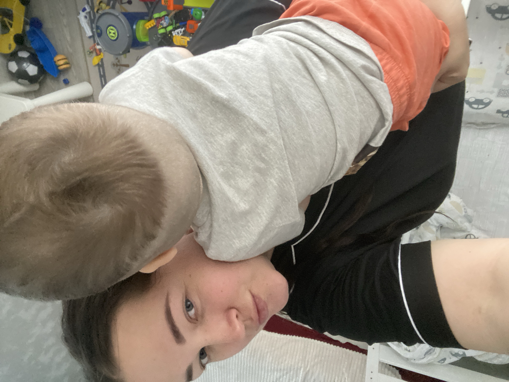 An indoor, portrait-oriented selfie captures a tender moment between an adult and a child in what appears to be a home in Sochi, Russia. The image is rotated, showing the adult's face on the left side of the frame.
The main subject is a woman with dark hair, blue eyes, and defined eyebrows, looking directly at the camera with a calm, soft expression. She is wearing a black top featuring white piping details around the collar or placket area. A baby or toddler is positioned directly on top of and behind her head, mostly obscured by the child's own body. Only the back of the child's head, with light brown hair, and upper body are clearly visible. The child is dressed in a light grey long-sleeved shirt and bright orange shorts.
The background reveals a brightly lit room, likely a child's play area or bedroom, with various toys scattered on a light-colored floor. Visible toys include a yellow car, a black and white soccer ball, a blue toy shovel, a small toy burger, and an assortment of colorful building blocks. A prominent grey and green toy drum or container with a yellow 'H' logo is also seen. To the right, part of a white bed frame or furniture is visible, along with a white blanket patterned with grey, abstract shapes, possibly cartoon car faces, suggesting children's bedding. The scene depicts a relaxed, everyday interaction, likely during daytime.