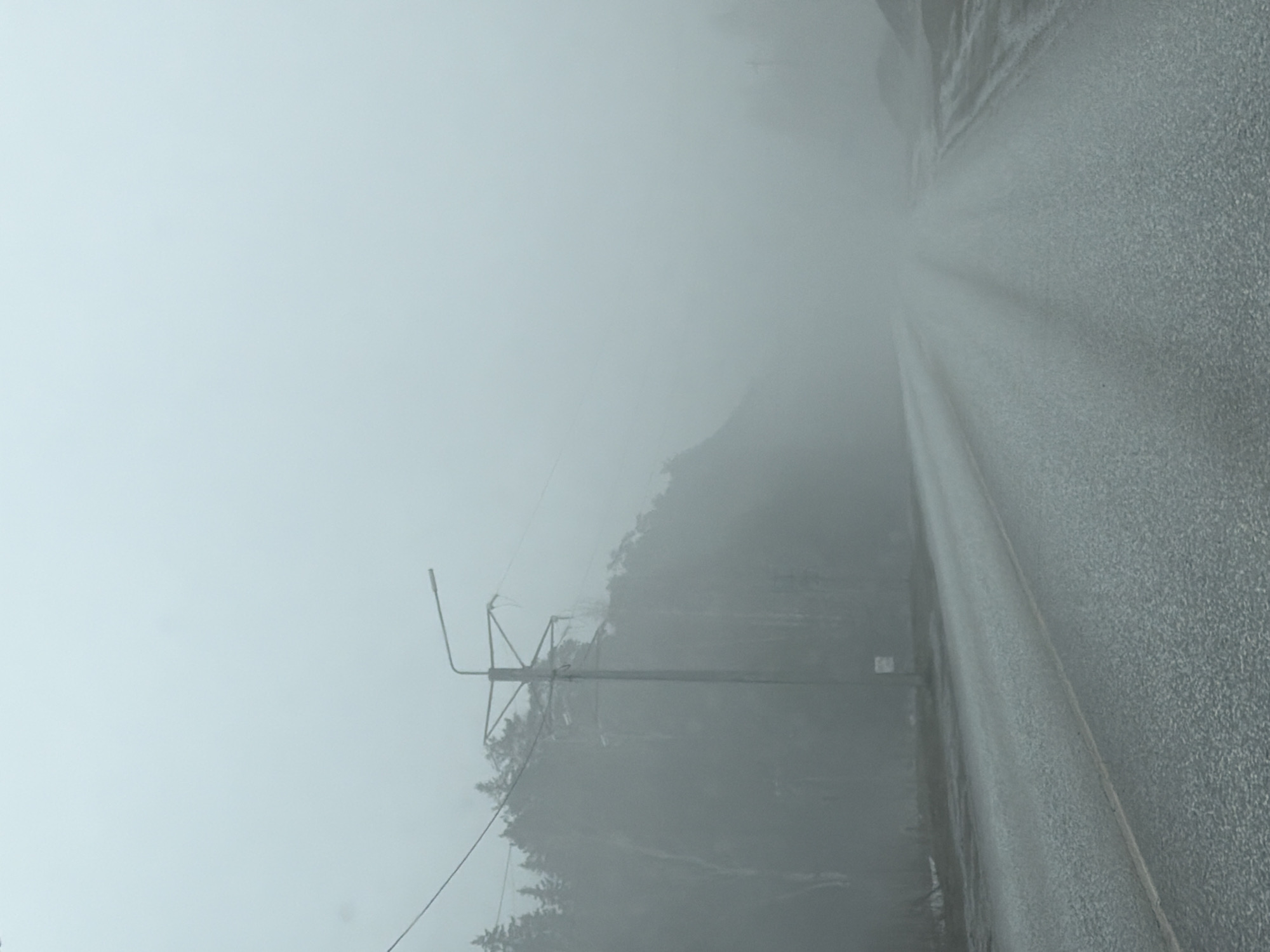 The image depicts a hazy scene in Chaikovsky, Russia. A road is visible on the right side, and a utility pole stands to the left, adjacent to what appears to be a tree-covered embankment or hillside. The weather is overcast or foggy, reducing visibility considerably. No people are visible.