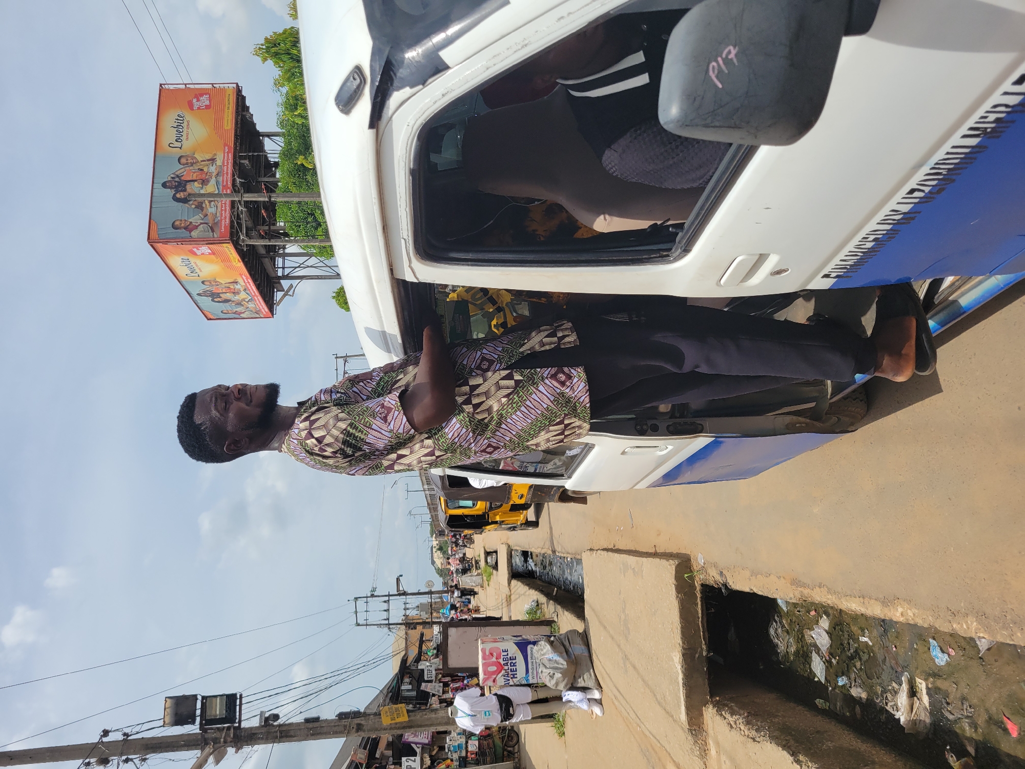 A man in a patterned shirt stands at the open door of a white commercial vehicle on a street in Egbagu, Nigeria. A billboard advertising "Lovebite" is visible in the background. A roadside drain with debris runs alongside the street. There are electrical poles and wires overhead, and pedestrians are seen further down the street. The area appears to be a bustling commercial area.