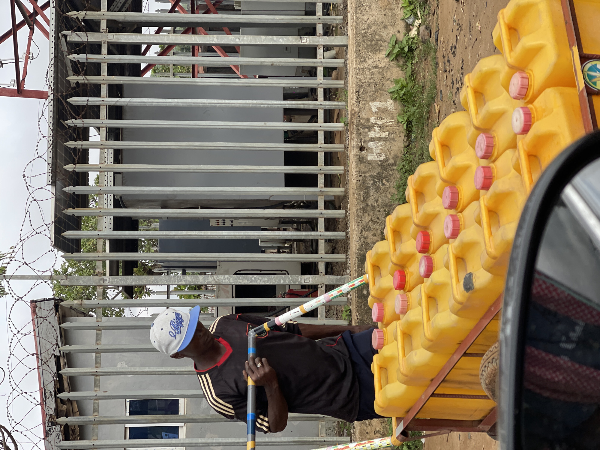 The image, taken in Abuja, Nigeria, shows a man wearing a black and red shirt with yellow stripes and a light blue and grey baseball cap. He is standing near a metal fence topped with barbed wire and spikes. In front of the fence, a cart or trailer is loaded with numerous yellow plastic jerrycans, each with a red or pink cap. The man is holding onto a pole, part of which is decorated with colorful patterns. The scene appears to be outdoors, with a cloudy sky visible above the fence. The ground is a mix of dirt and concrete. The overall impression is of an industrial or commercial area, possibly a storage or distribution point for the jerrycans. No specific time of day or strong emotions are discernible, but the overcast sky suggests it might be a humid day.