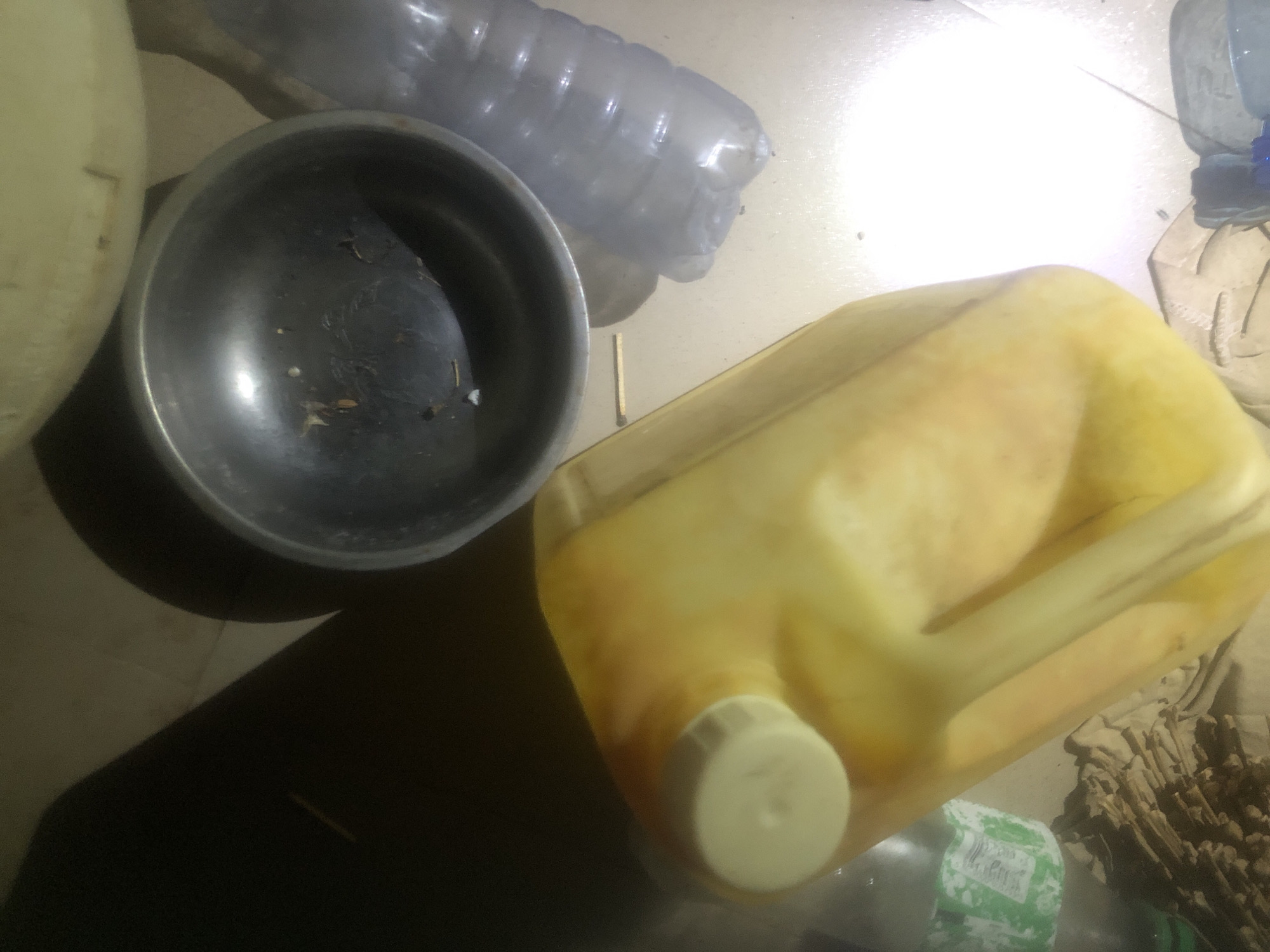 The image depicts a cluttered indoor scene in Wukari, Nigeria. In the foreground, a large, yellow plastic jerrycan with a white cap is prominently displayed. To its right, a partially filled, clear plastic water bottle is visible. Behind these items, a dark metal bowl containing some debris sits next to another clear plastic bottle. A portion of a larger, light-colored container is visible at the top of the frame. In the lower left, a stack of what appears to be dried plant material or kindling is present, along with a green and white labeled bottle partially obscured. The lighting is uneven, with a bright spotlight effect in the lower right quadrant, suggesting artificial illumination. The floor appears to be tiled. The overall impression is of a storage area or a domestic space with various items gathered together. There are no people or animals visible in the image.