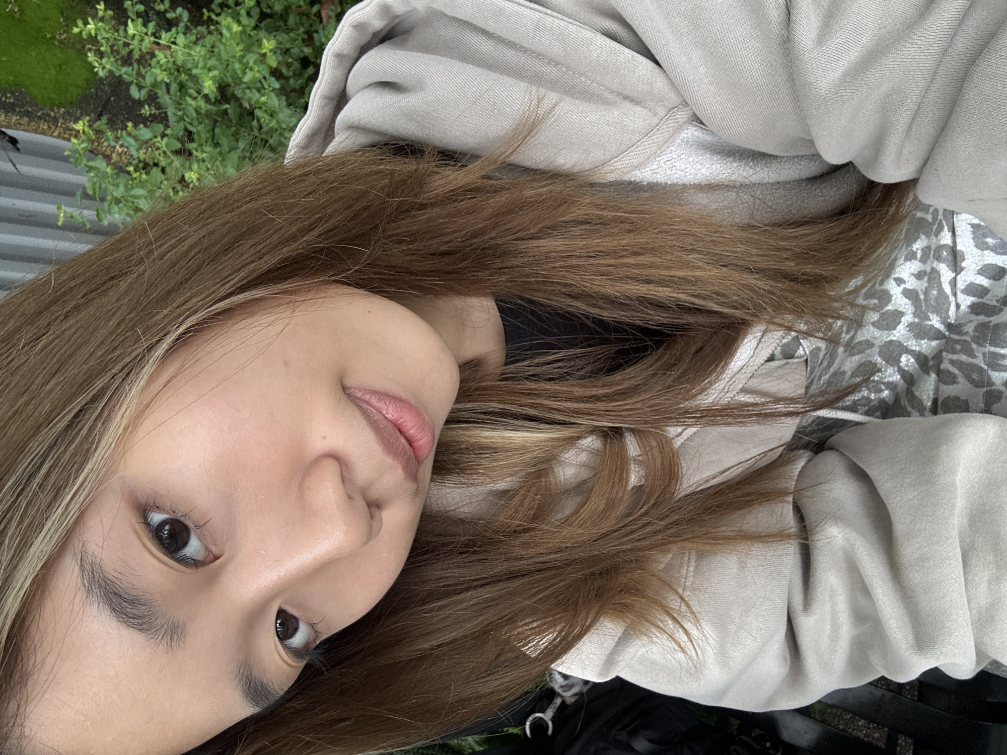 A young woman with brown hair, wearing a light brown hooded sweatshirt, is outdoors. The setting appears to be a park or garden, with greenery visible in the background. The woman is looking towards the camera, with a slight smile. The location in Yuzhong District, China is not discernible from the image.
