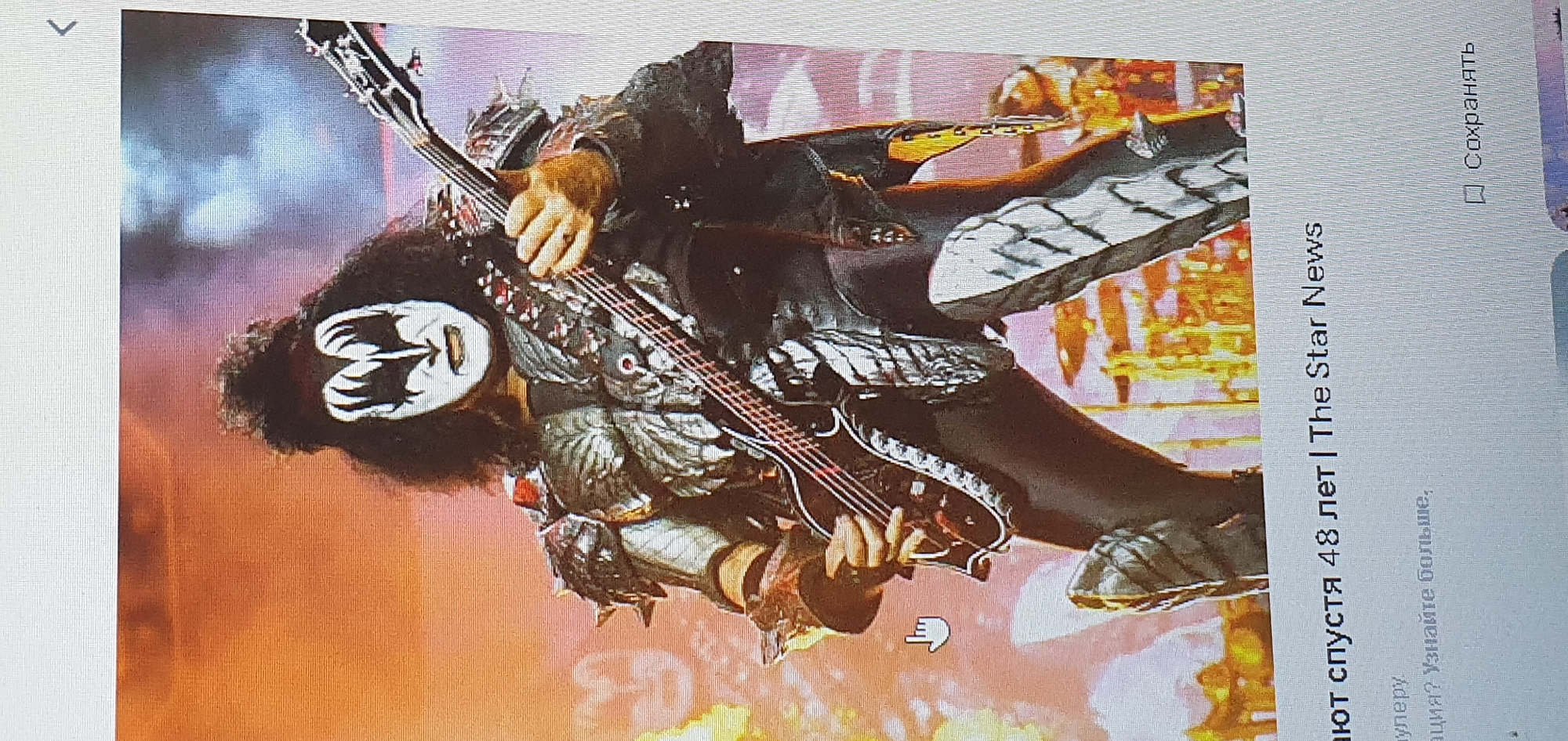 The image shows a close-up of a musician, identified as Gene Simmons of the band KISS, performing on stage. He is wearing a theatrical costume with white face paint, black makeup around his eyes, and a black wig. He is playing a black bass guitar with red strings. His costume includes elaborate armor-like shoulder pads and leg coverings. The background is blurred and illuminated with warm orange and purple lights, suggesting a concert setting. Below the image, text in Russian and English is visible, including "The Star News" and "Сохранять" (Save). The image does not contain any information or visual cues related to the city of Bat Yam, Israel.