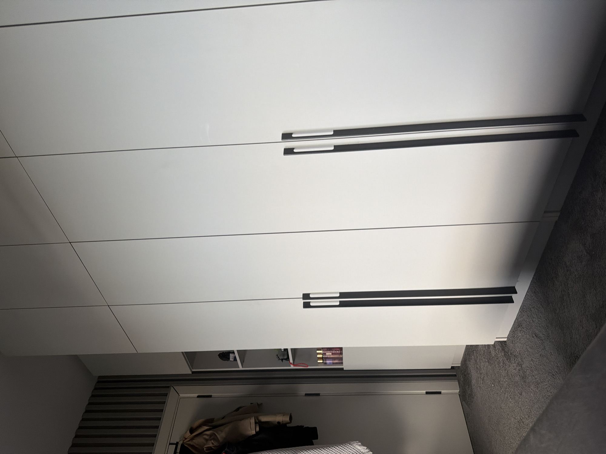 The image shows a close-up view of a modern wardrobe with clean, white doors and minimalist black handles. The wardrobe appears to be made of smooth, possibly lacquered, material. There are horizontal lines that divide the doors into sections. In the lower section of the wardrobe, an open shelf is visible, containing several bottles of what appear to be perfume or body spray, some with pink liquid. There is also a black object, possibly a speaker or a small electronic device, and a red cord. Below this shelf, more clothing is visible, including a tan jacket and other garments. The floor in front of the wardrobe is covered with a dark grey carpet. The lighting suggests it is indoors, likely during the daytime. The scene is tidy and suggests a domestic setting, possibly a bedroom or dressing area. The image is taken from a slightly low angle, looking up at the wardrobe. The location is Ruse, Bulgaria. There is no visible text in the image.