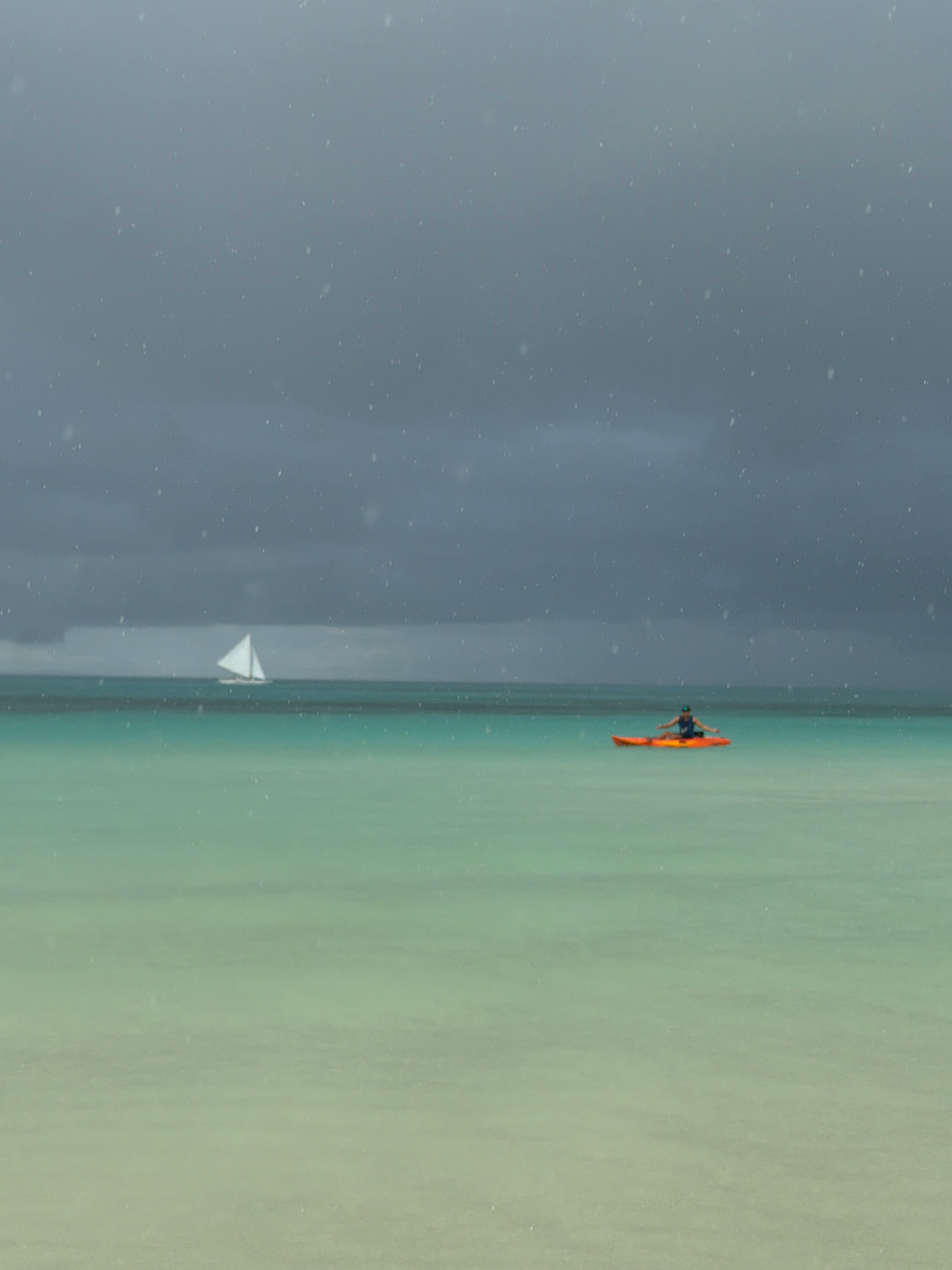 In the ocean off Malay, Philippines, under a grey, overcast sky with visible rain or mist, a person is kayaking in an orange kayak. The person is seated and appears to be paddling. In the distance, a sailboat with a white sail is visible on the horizon. The water is a gradient of turquoise and pale green, with lighter sand visible underneath the surface in the foreground.