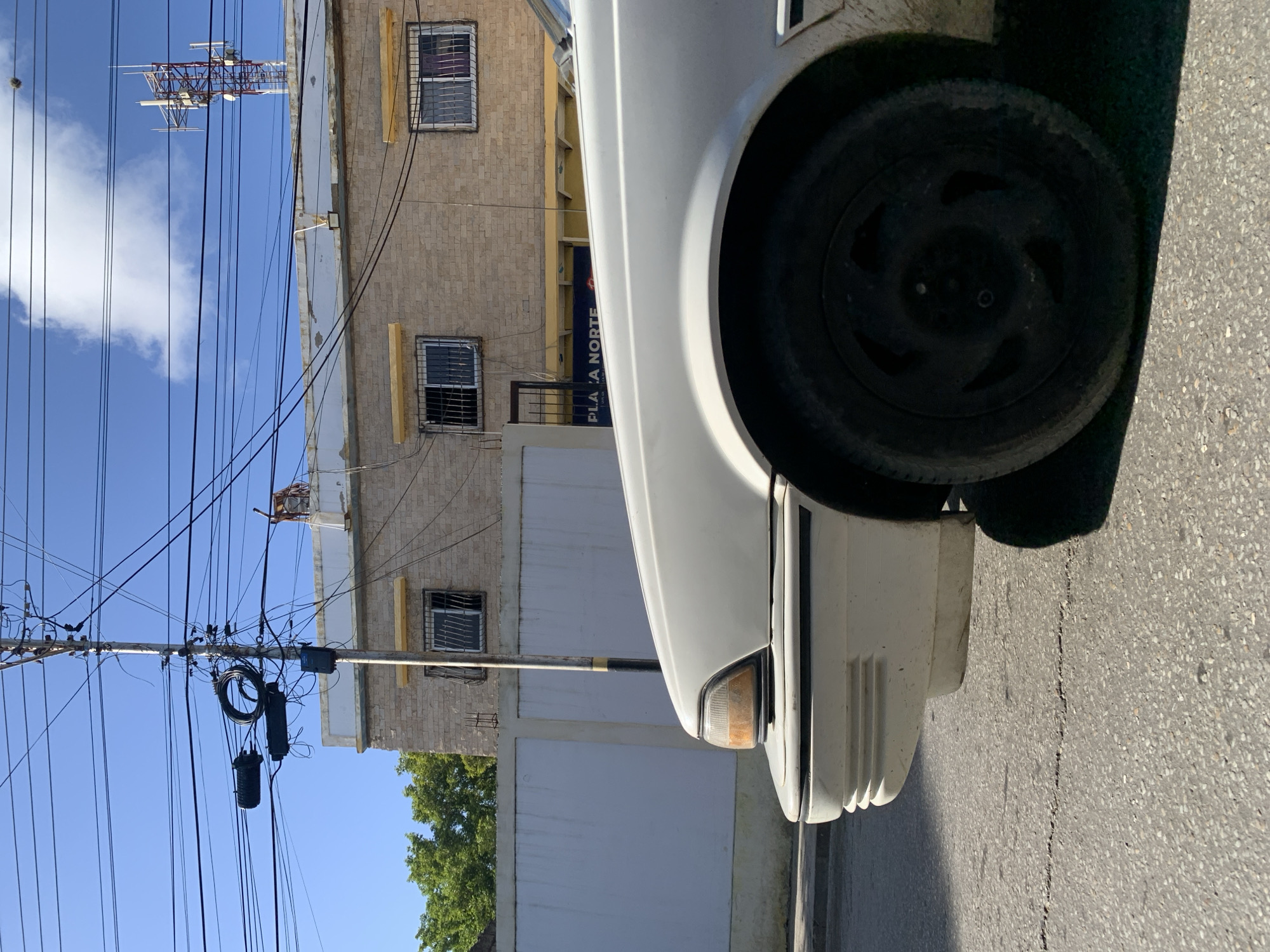 The image shows the front passenger side of a white car parked on a street in Santa Ana, Venezuela. The car's headlight and wheel are visible. In the background, there is a building with barred windows and a sign that reads "PLATA NORTE". A utility pole with numerous wires and a cell tower can be seen against a blue sky with some clouds. The street appears to be asphalt and shows some cracks. Trees are visible to the left of the building. The lighting suggests it is daytime.