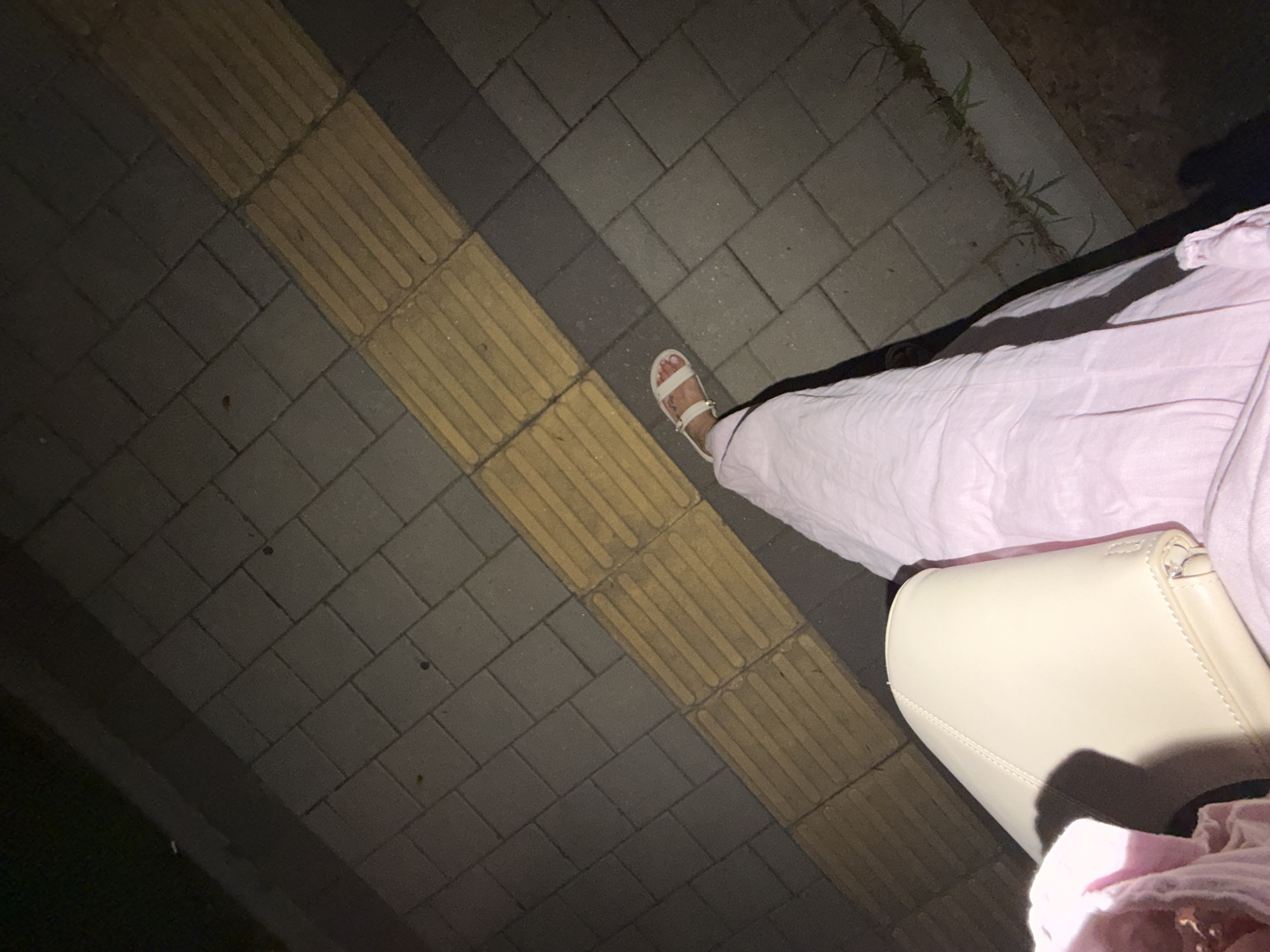 The image displays a close-up view of a person's legs and a handbag, situated on a patterned pavement. The pavement consists of grey rectangular tiles arranged in a repeating pattern, interspersed with a diagonal strip of yellow tactile paving bricks. The yellow bricks have raised linear patterns, typical of those found on sidewalks to assist visually impaired pedestrians. The lighting suggests it is nighttime, with artificial light casting shadows. The person is wearing light pink pants and white sandals. A light-colored handbag is visible, resting on the person's lap. The environment appears to be an outdoor urban setting, possibly a sidewalk or pedestrian area in the city of Pravets, Bulgaria. The scene is captured from a high-angle perspective, looking downwards.