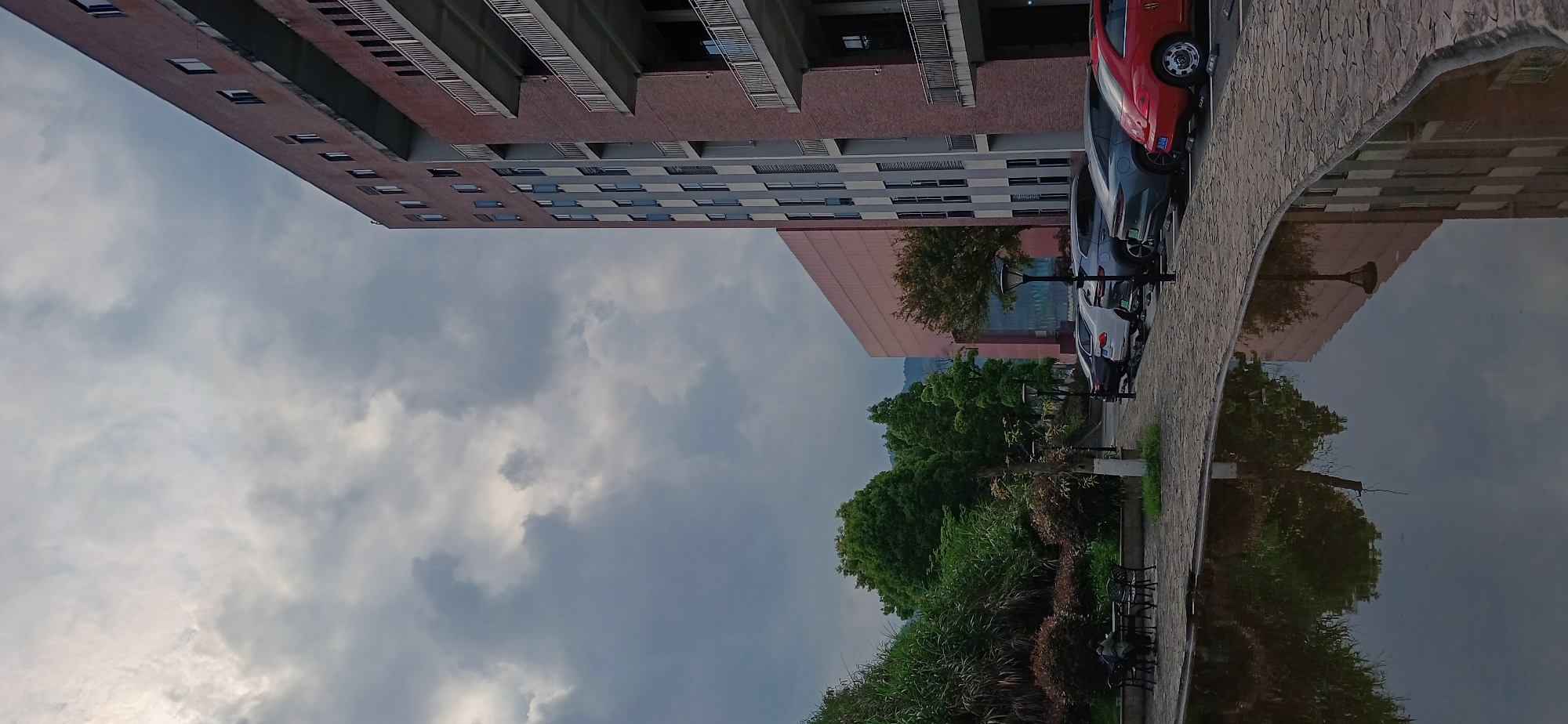 The image depicts a reflection of a cityscape in Chongqing High-tech Zone, China. A multi-story brick building with multiple balconies and windows is visible along the left side of the image and is reflected in a dark, mirror-like surface. A row of parked cars, including a red one, lines the edge of a stone-paved walkway. Green trees and shrubbery are also reflected in the water. An overcast sky is above.