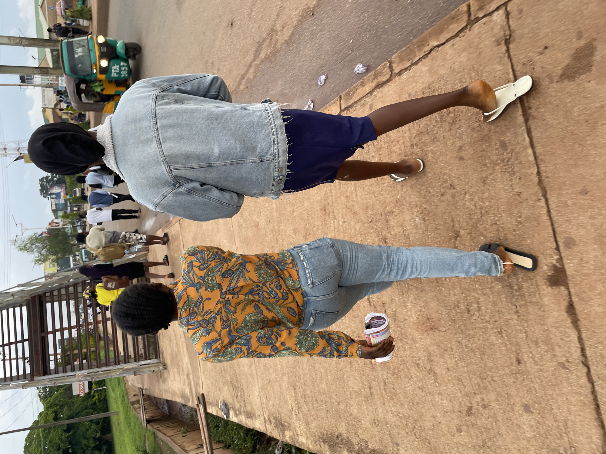 The image shows a street scene in Jos, Nigeria. Two individuals are walking away from the camera on a sidewalk. The person in the foreground on the left is wearing a colorful patterned long-sleeved shirt and blue jeans. They are carrying a rolled-up item in their right hand. The person on the right is wearing a denim jacket, a dark blue skirt, and white flip-flops. A green and yellow tricycle taxi is visible on the street to the right, along with other vehicles and pedestrians in the background. An overpass structure is present on the left side of the image, with a drainage ditch running alongside the sidewalk. The overall environment appears to be an urban setting with moderate foot and vehicle traffic.