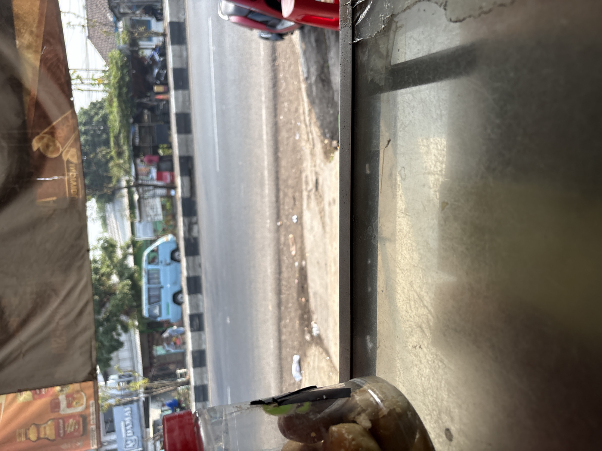The image captures a street view through a window, likely from a shop or restaurant, in Palimanan, Indonesia. In the foreground, a clear plastic container filled with round, light-brown objects, possibly food items, is visible on the left. The container has a red lid and a black sticker on its side. Behind the container, a metallic surface reflects the scene. The middle ground shows a paved road with a black and white striped curb. A blue van is driving on the road, and several people are seen near stalls or shops on the other side. The background features buildings, trees, and a cloudy sky. A red scooter and a red plastic chair are parked on the side of the road. The overall impression is of a bustling street scene in a tropical or subtropical urban environment.