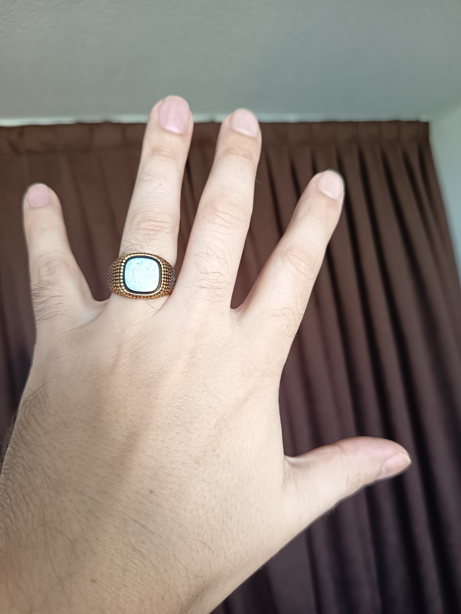 A person's hand wearing a square, light blue stone ring with a textured gold band is held up against a background of brown curtains. The hand is positioned with the palm facing the camera, and the fingers are spread open, with the thumb bent. The ring is on the ring finger of the left hand. The scene is indoors, likely in a room with natural lighting. The image is focused on the hand and the ring, with the background blurred. There are no visible identifying marks of the city of Izmail, Ukraine.