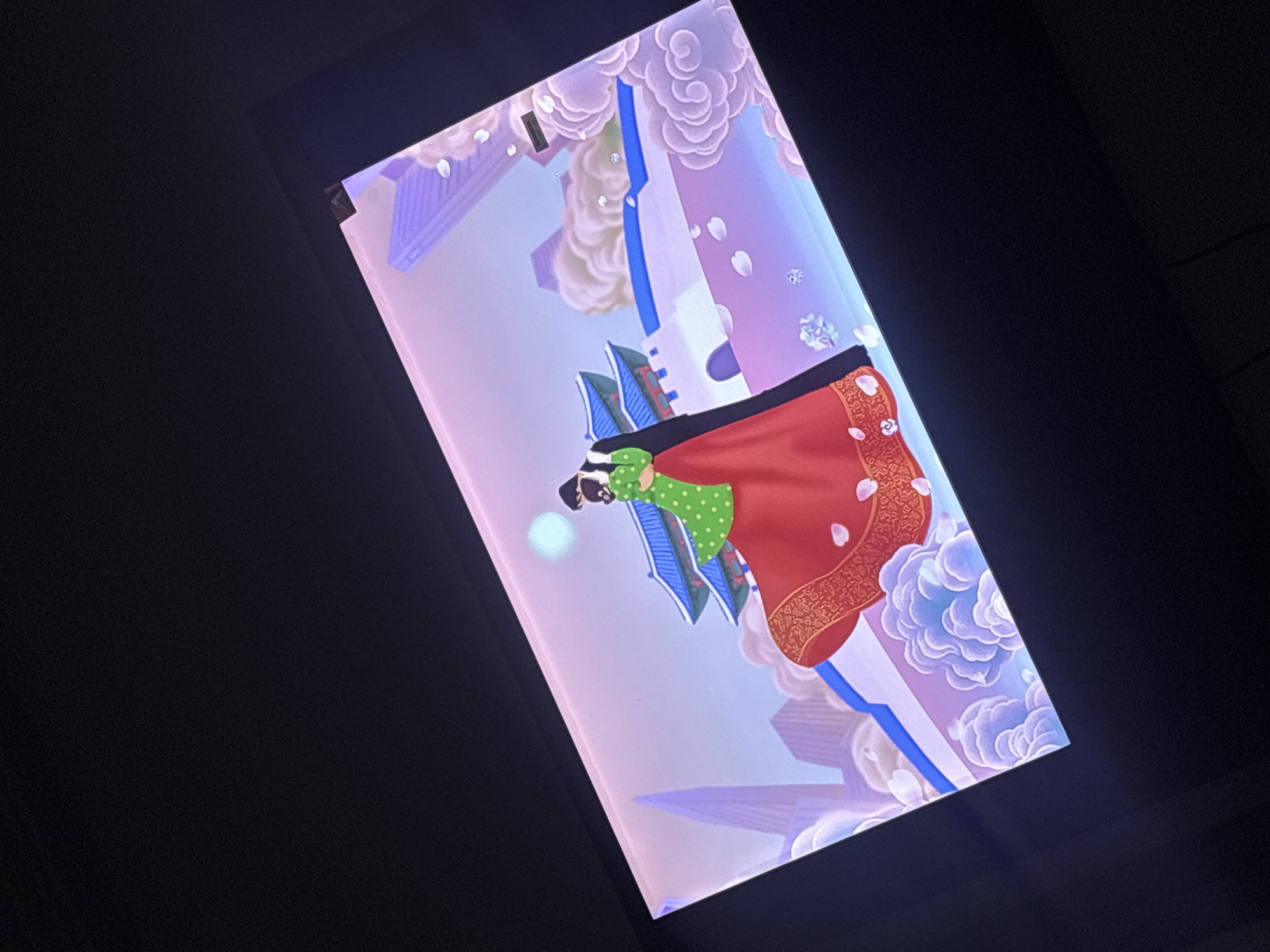 A digital screen displays an illustration. The illustration features an individual in a green dotted dress and a red and gold patterned shawl, positioned against a backdrop of stylized clouds and architecture. The color palette is predominantly pastel, with shades of purple, white, and blue. The screen is framed by a dark, almost black, surround. The specific location of the image or screen in Port Harcourt, Nigeria, is not discernible.
