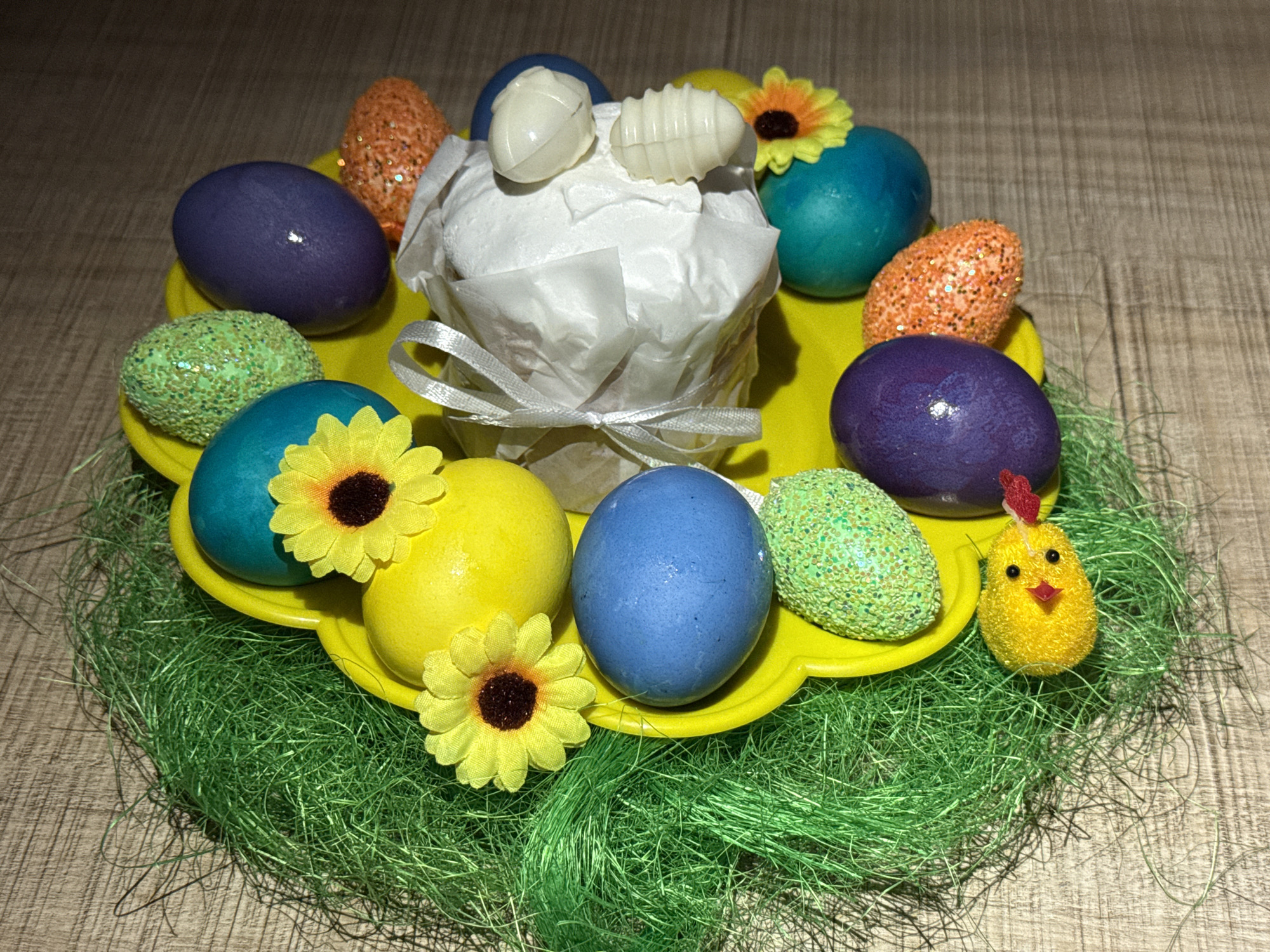The image shows an Easter display. Centered is a small cake wrapped in white paper and tied with a silver ribbon. On top are two white chocolate decorations. The cake sits on a yellow plastic plate surrounded by Easter eggs in various colors: purple, teal, orange with glitter, yellow, blue, and green with glitter. Two yellow sunflowers decorate the plate. The plate rests on a bed of green artificial grass. A yellow chick with a red comb stands beside the plate. The background is a light brown wooden surface. The scene appears to be indoors, likely taken in the city of Chaikovsky, Russia given the instructions.