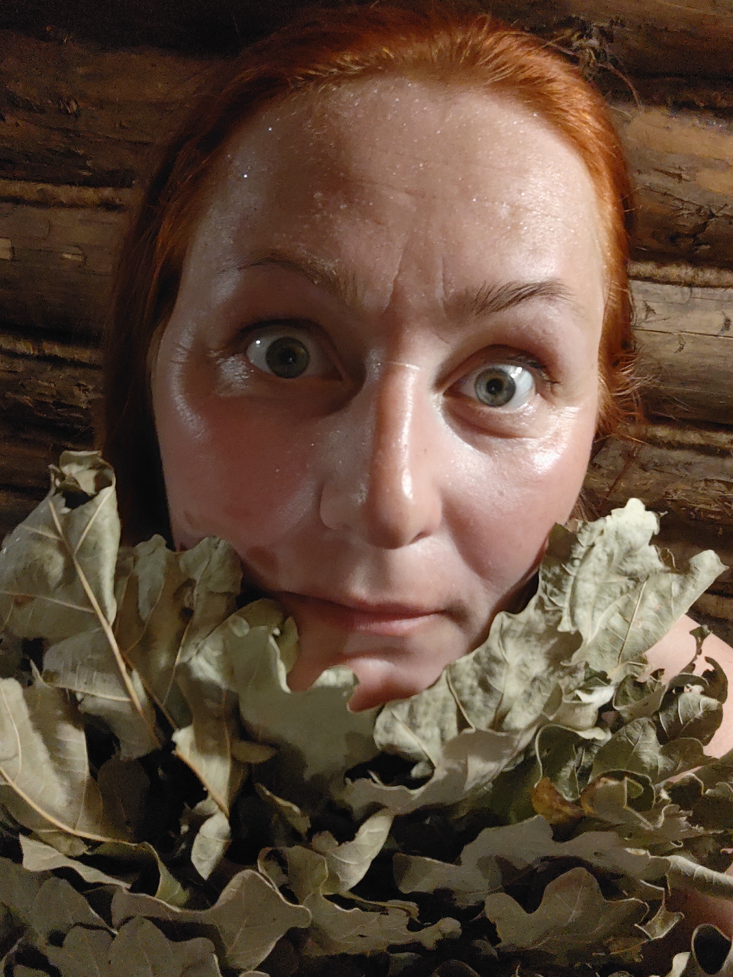 A close-up photograph depicts a woman with red hair and a fair complexion, positioned against a background of interlocking wooden logs, possibly in Фоки, Russia. Her face appears flushed and glistening, possibly due to sweat. She is holding a bundle of dried oak leaves, which are obscuring part of her chest and neck. Her eyes are wide and expressive.