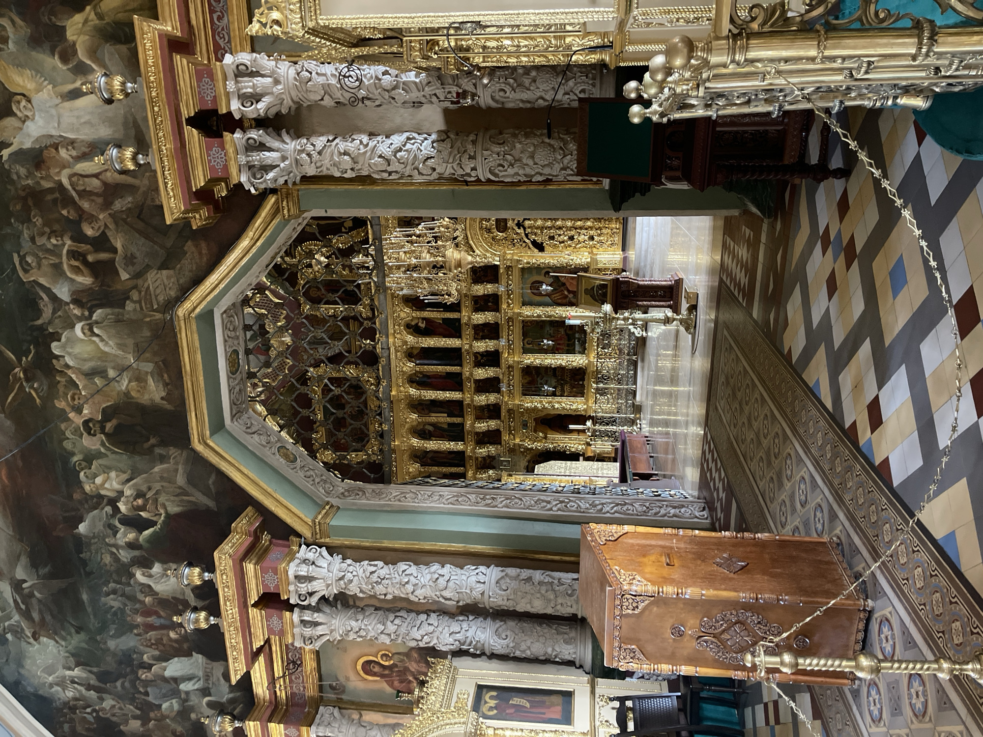 The image depicts the interior of the Dormition Cathedral in Sergiyev Posad, Russia. The scene is characterized by opulent religious decoration. In the foreground, a wooden pulpit with intricate carvings stands to the left, and to the right, a gilded metal railing with a turquoise fabric barrier. The floor is tiled with a geometric pattern of beige, brown, red, and blue squares and rhombuses.
A long, patterned carpet runner extends down the center aisle. Beyond the foreground, an archway leads into the main sanctuary, revealing a highly ornate iconostasis, a wall of icons, heavily adorned with gold. This iconostasis is a focal point, featuring multiple tiers of icons enclosed in gilded frames and decorative elements. A large, elaborate chandelier hangs in the sanctuary.
The walls and ceiling are adorned with a vast mural, likely depicting biblical scenes or religious figures. The mural is rendered in a classical style with a multitude of people and celestial beings. The architectural elements, such as the columns with elaborate carvings and gilded capitals, contribute to the grandeur of the space. The overall impression is one of immense wealth and spiritual significance, typical of a historic Russian Orthodox cathedral.