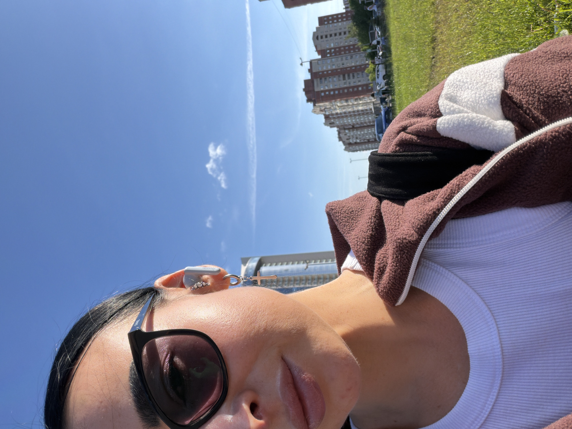 A brightly lit outdoor selfie, rotated 90 degrees clockwise from its natural orientation, captures a young woman and an urban landscape. The primary subject is a woman, visible from the mid-chest up, with dark hair and dark sunglasses obscuring most of her eyes. She wears a white ribbed tank top under a cozy brown and white fluffy hooded jacket with a white zipper. Her right ear, adorned with small hoop earrings, also holds a white ear-worn device, possibly an earbud or a charm holder, from which a small silver cross charm dangles. Faint text, likely a brand name such as "AWEI," is visible on this device.
The background, set in what would be the right and upper portions of a correctly oriented image, features a vibrant green grassy area. Beyond the grass, a row of multi-story residential buildings with red and lighter-colored facades can be seen, bordering what appears to be a street or parking lot with parked cars. The dominant feature of the sky, occupying the upper left, is a clear, bright blue expanse with a few wispy white clouds and a long, thin airplane contrail stretching across it.
The activity taking place is a casual outdoor selfie, suggesting the person is enjoying the pleasant weather. The bright sunlight indicates it is midday or early afternoon. The setting is an urban environment with a mix of residential buildings and green spaces, consistent with Saint Petersburg, Russia. The overall mood is relaxed and casual.